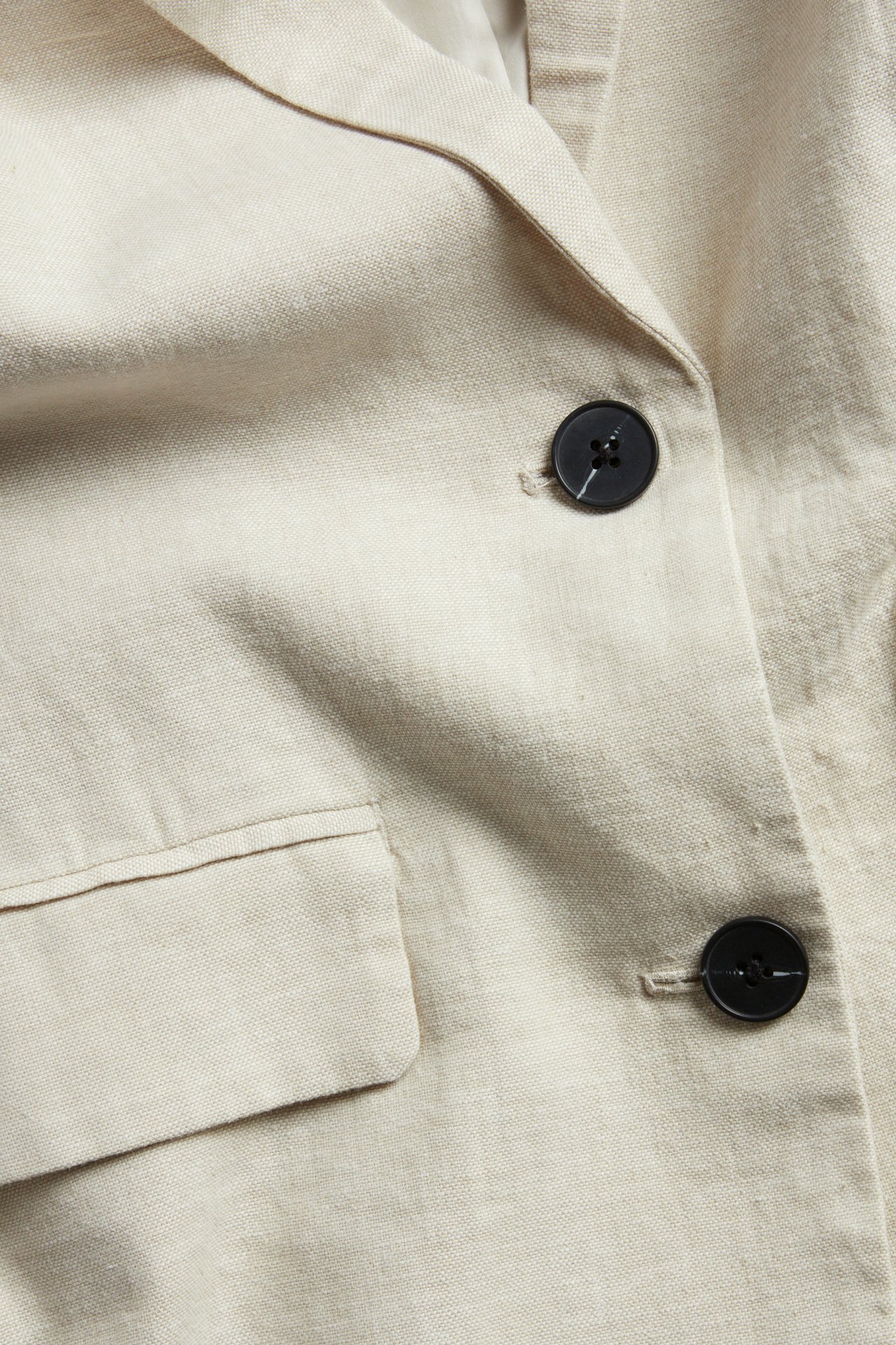 Zbliżenie beżowej marynarki z mieszanki lnu z czarnymi guzikami i klapką kieszeni.