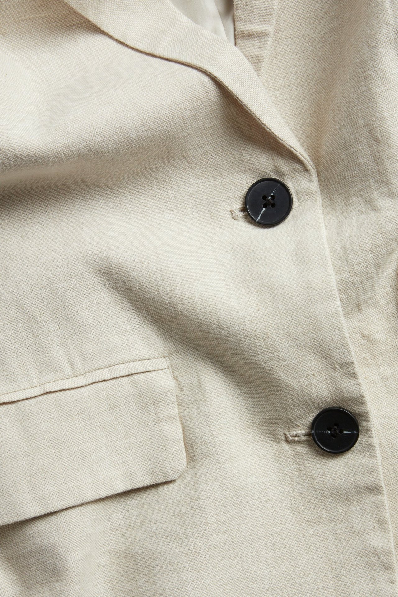 Zbliżenie beżowej marynarki z mieszanki lnu z czarnymi guzikami i klapką kieszeni.