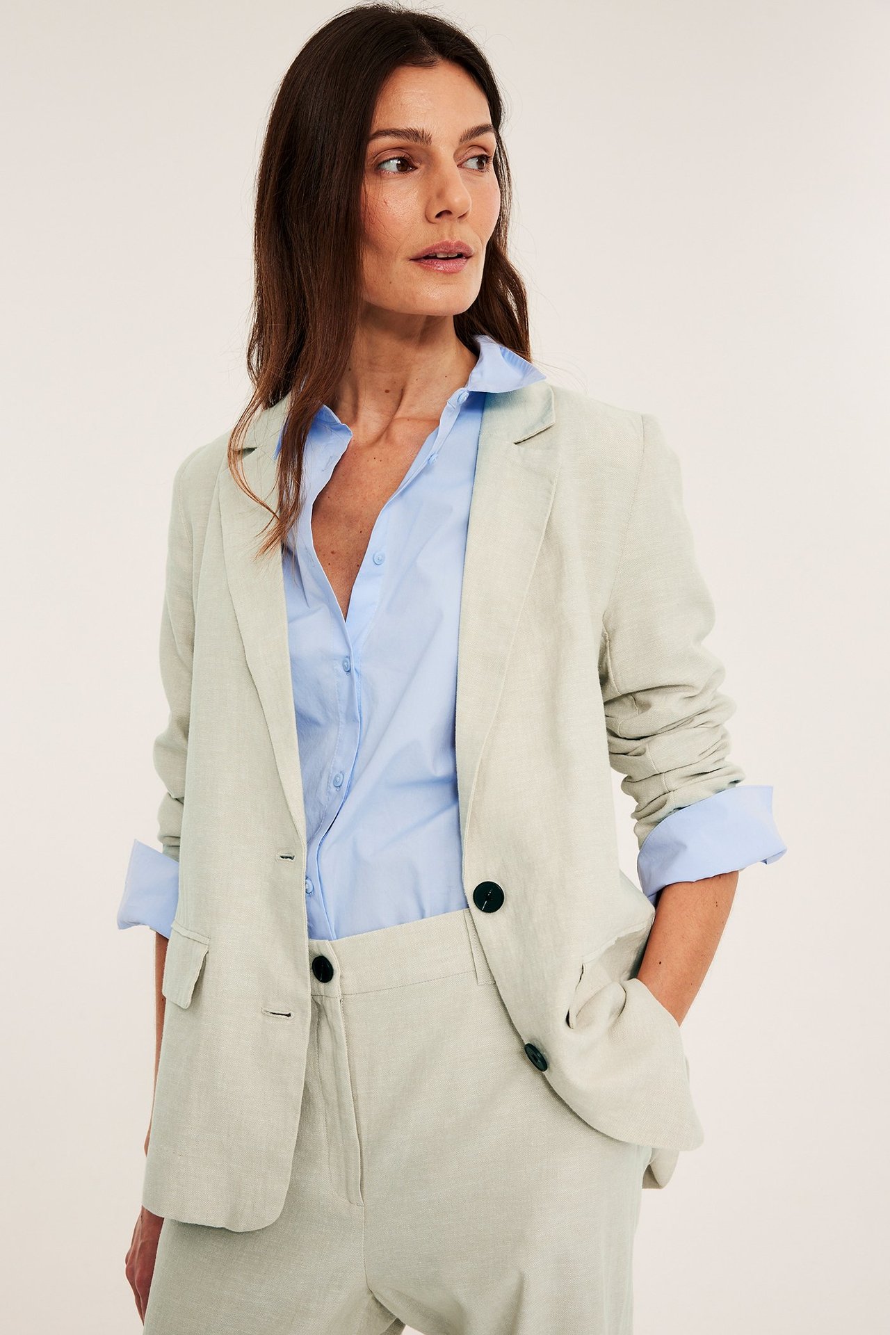 Kvinne i åpen melert beige linblanding-blazer over lyseblå skjorte og matchende bukser.