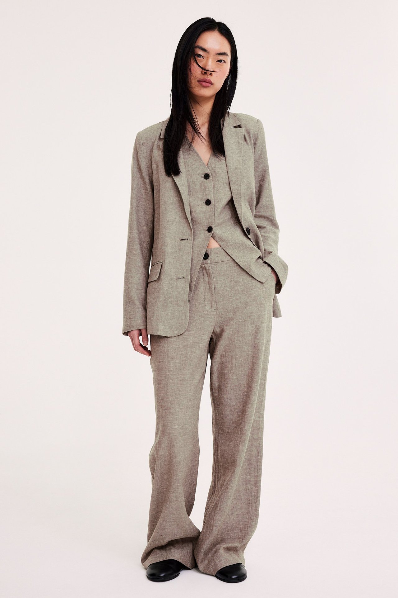 Naisen edestä päin, yllään kirjava vaaleanruskea pellavasekoitteinen bleiseri, liivi ja leveälahkeiset housut.