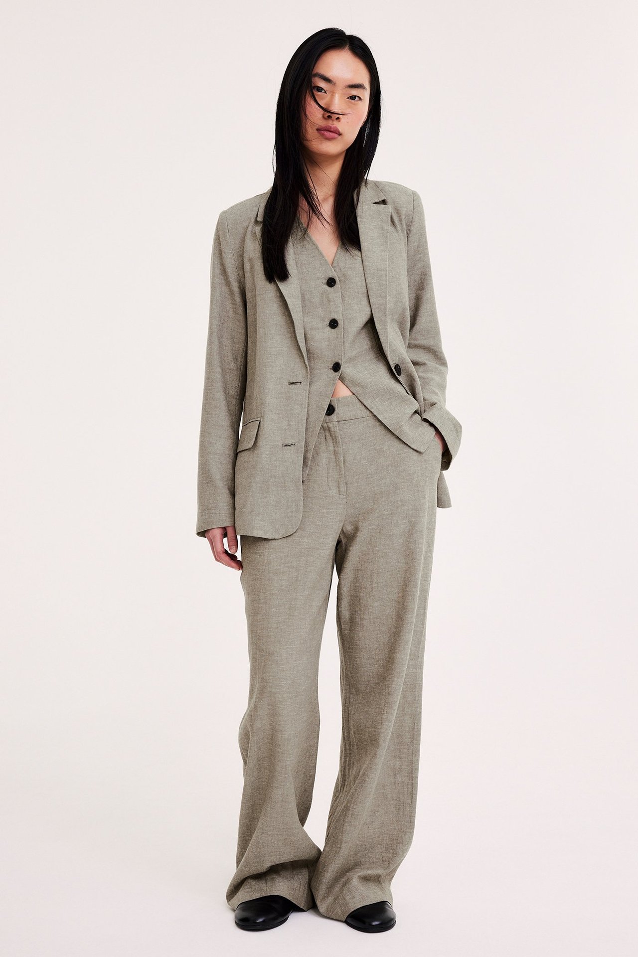 Framifrån: Kvinna i brunmelerad linneblazer, matchande väst, vida byxor och svarta platta skor.