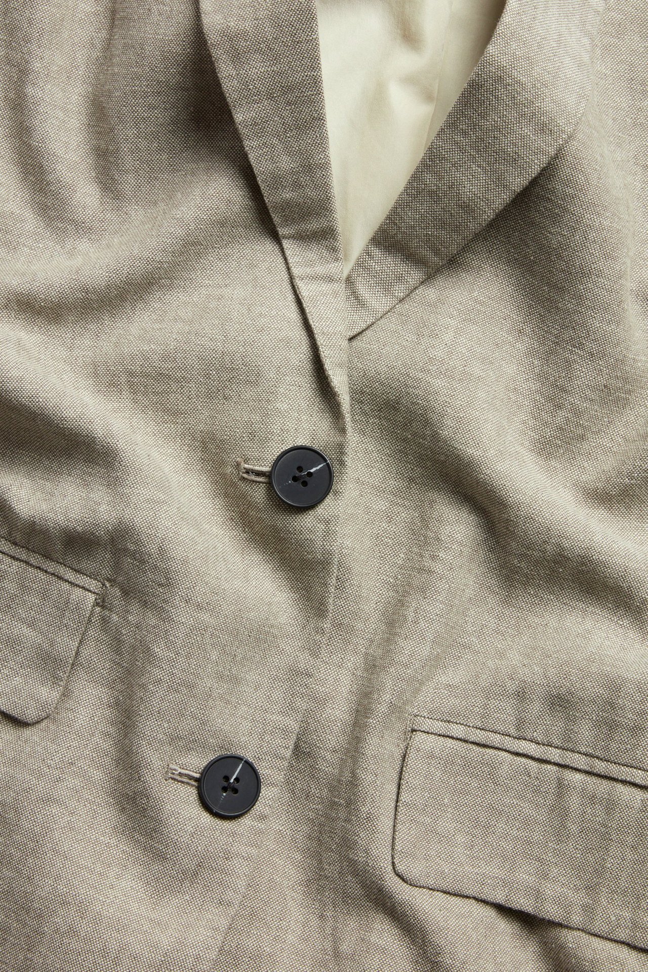 Zbliżenie na beżową melanżową marynarkę z mieszanki lnu, z wyciętymi klapami i dwoma ciemnymi guzikami.