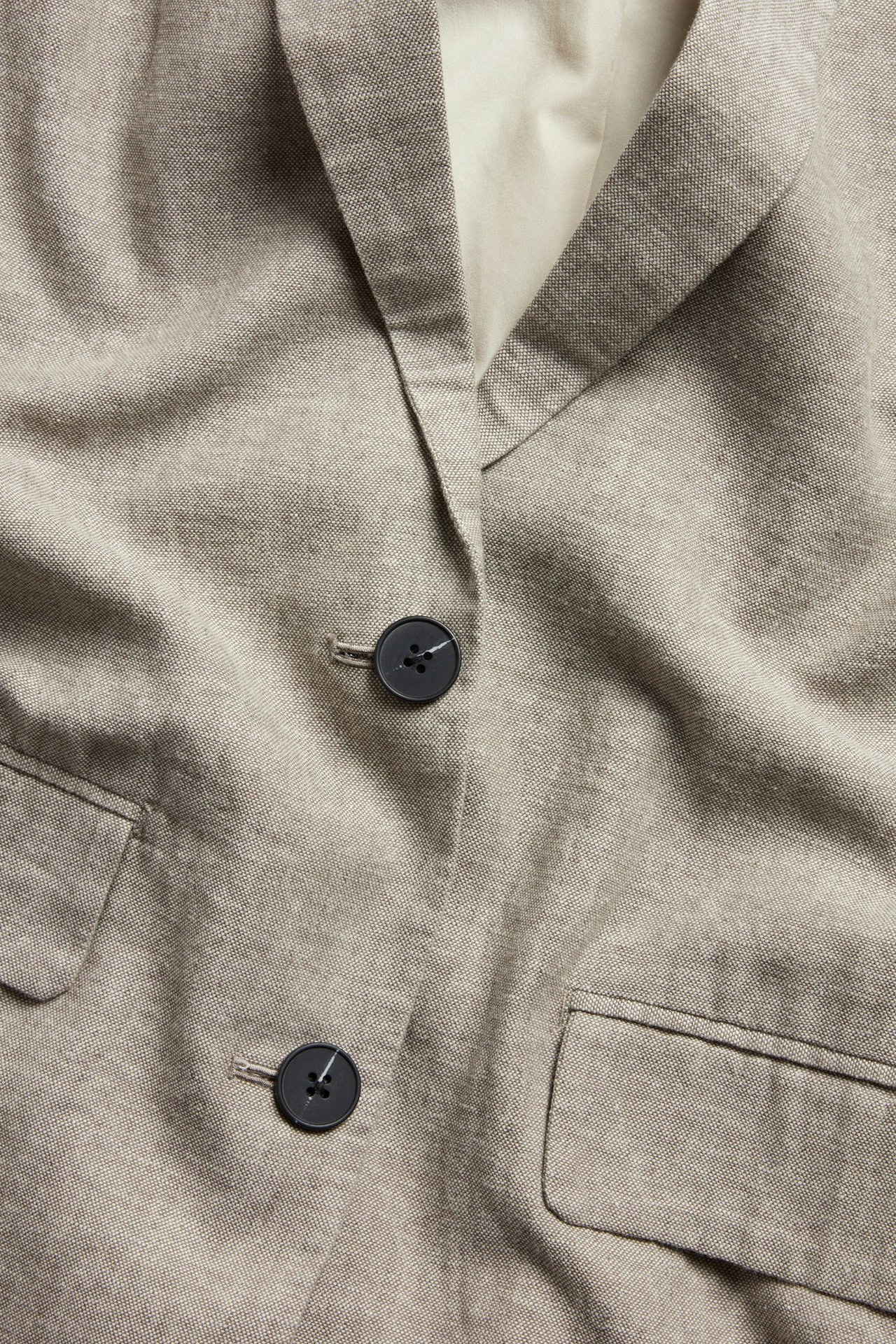 Zbliżenie, przód brązowej melanżowej marynarki lnianej z dwoma czarnymi guzikami i kieszeniami z klapą.