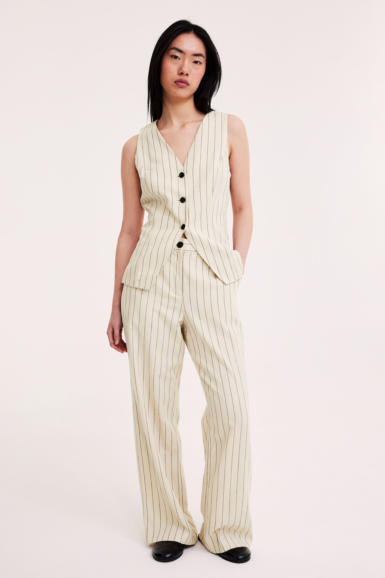 Edestä: Nainen yllään luonnonvalkoinen raidallinen pellavasekoiteliivi mustilla napeilla ja yhteensopivat leveälahkeiset housut.