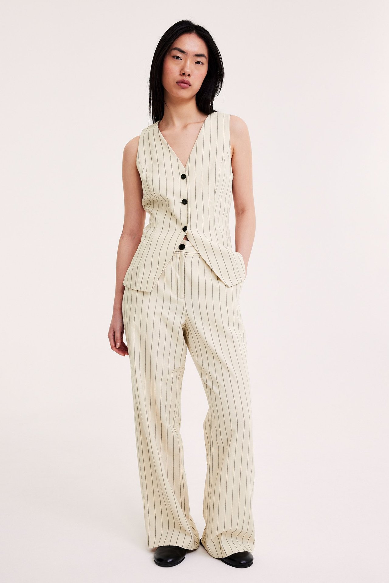 Edestä: Nainen yllään luonnonvalkoinen raidallinen pellavasekoiteliivi mustilla napeilla ja yhteensopivat leveälahkeiset housut.