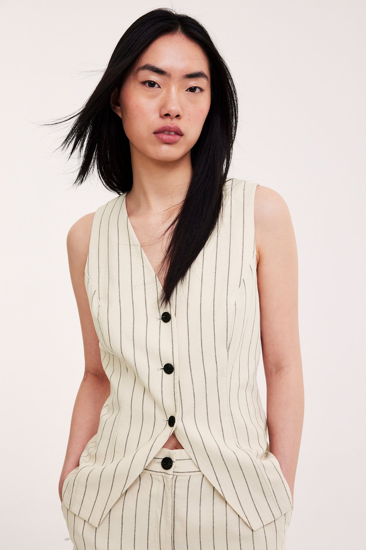 Etunäkymä naisesta, yllään offwhite raidallinen pellavasekoite liivi mustilla napeilla ja siihen sopivat housut.