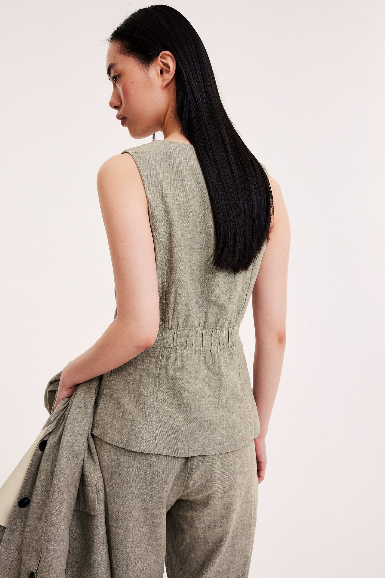 Takasivuprofiili naisesta yllään kapea, kirjava vaaleanruskea/beige pellavasekoitevät, joustava selkä ja housut, pitäen bleiseriä.