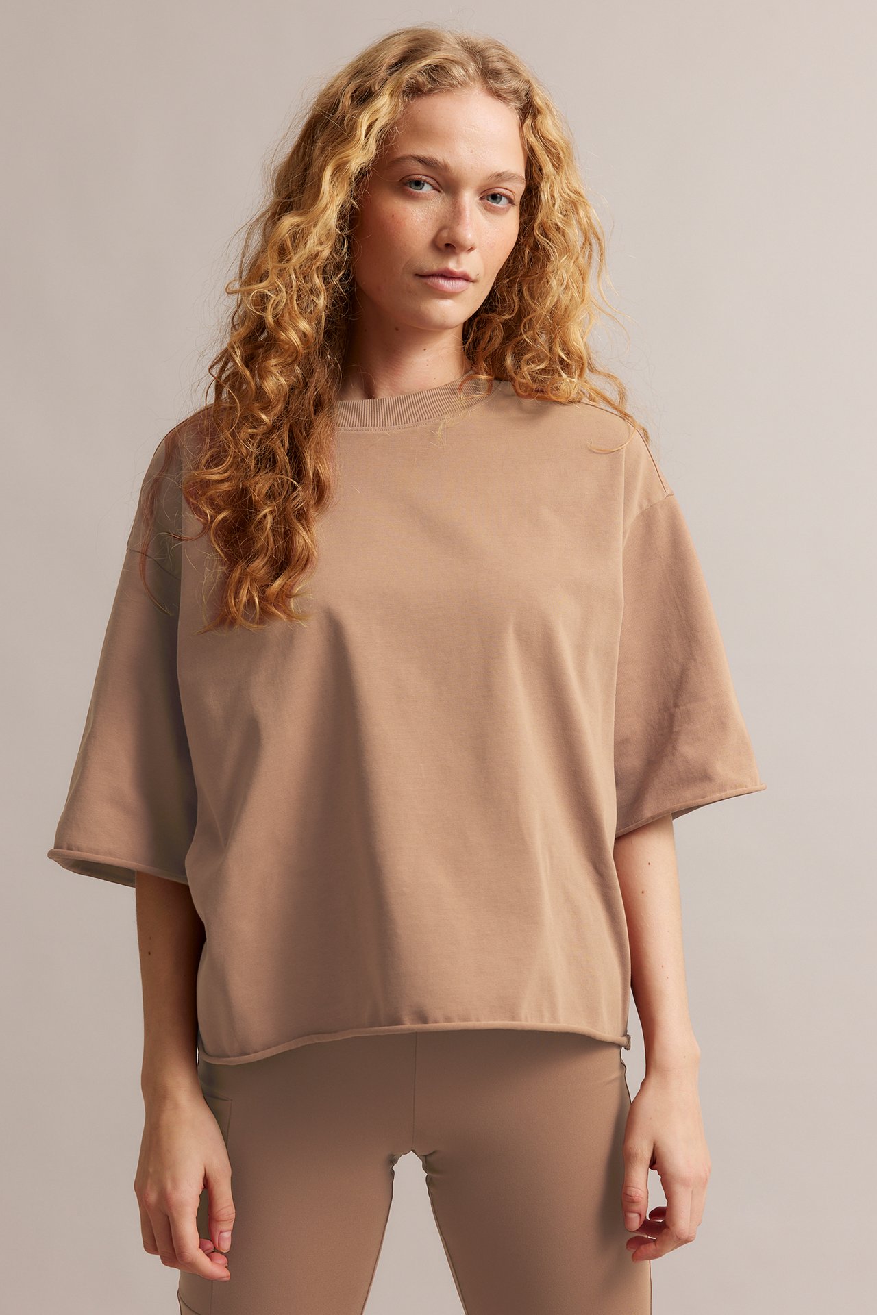 Forfra, kvinne i en avslappet brun bomulls T-skjorte med råe kanter og matchende brune leggings.
