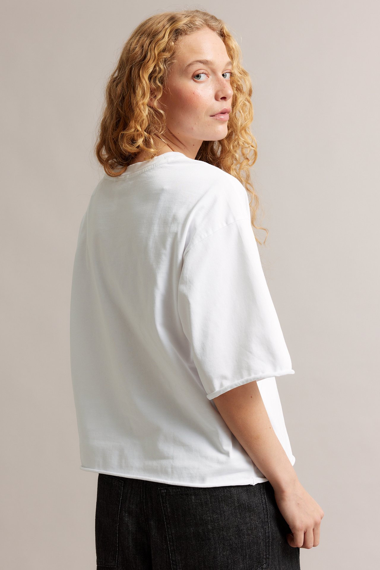 Kvinne i løstsittende hvit T-skjorte av bomull med rå kanter og mørke bukser, sett bakfra/fra siden.