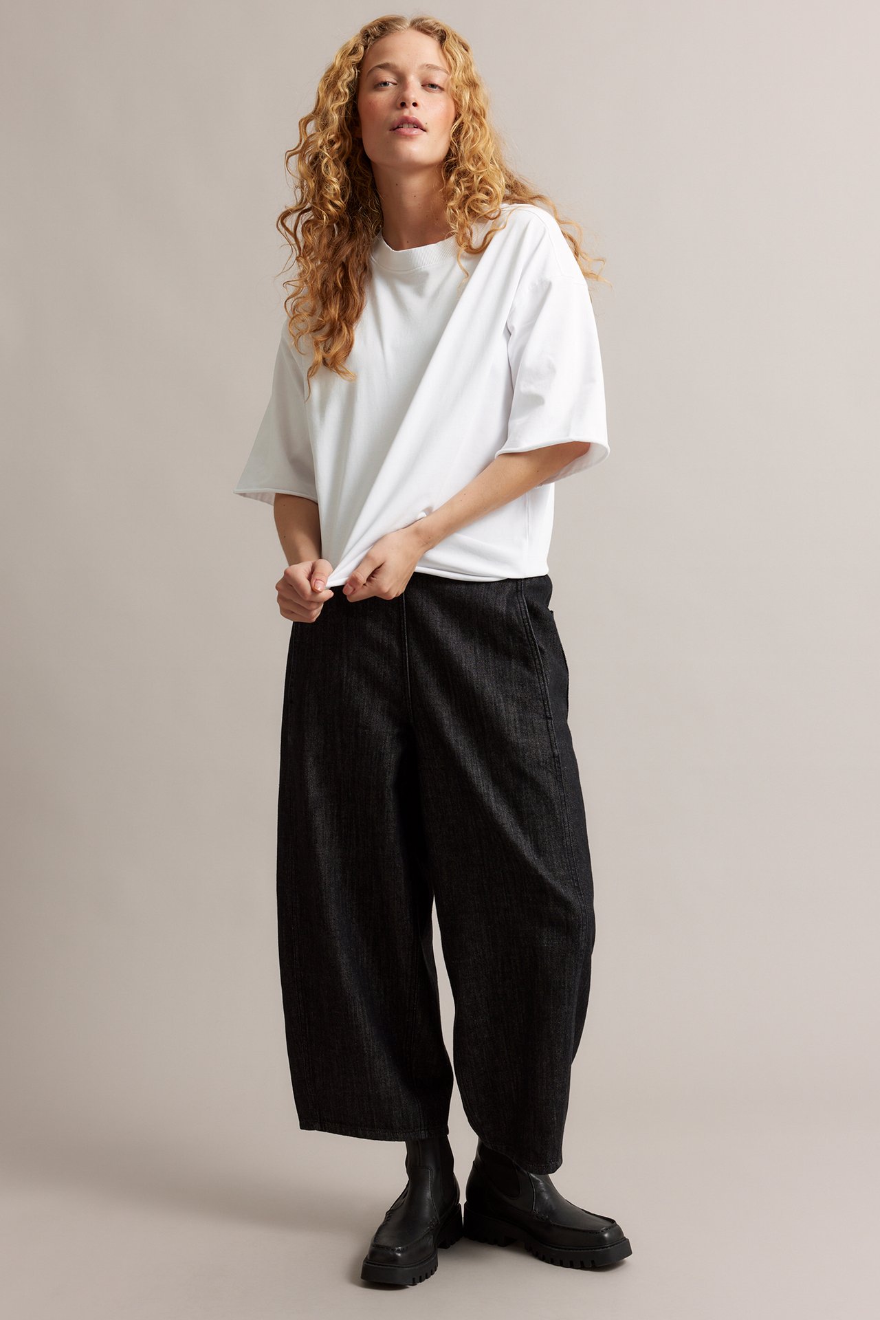 Frontbilde: Kvinne i hvit oversized T-skjorte med råkanter, mørke vide bukser og svarte chunky ankelstøvletter.