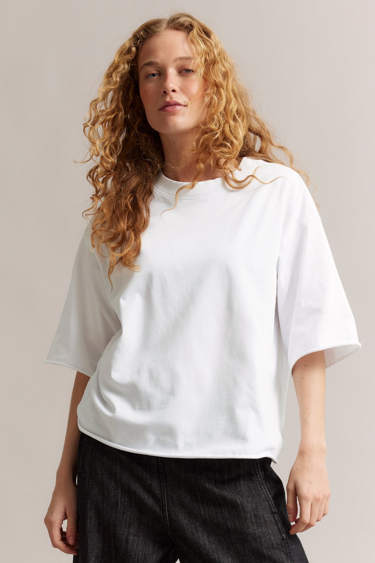 Forfra: kvinne i en hvit, løstsittende t-skjorte i bomull med råe kanter, kombinert med mørke bukser.