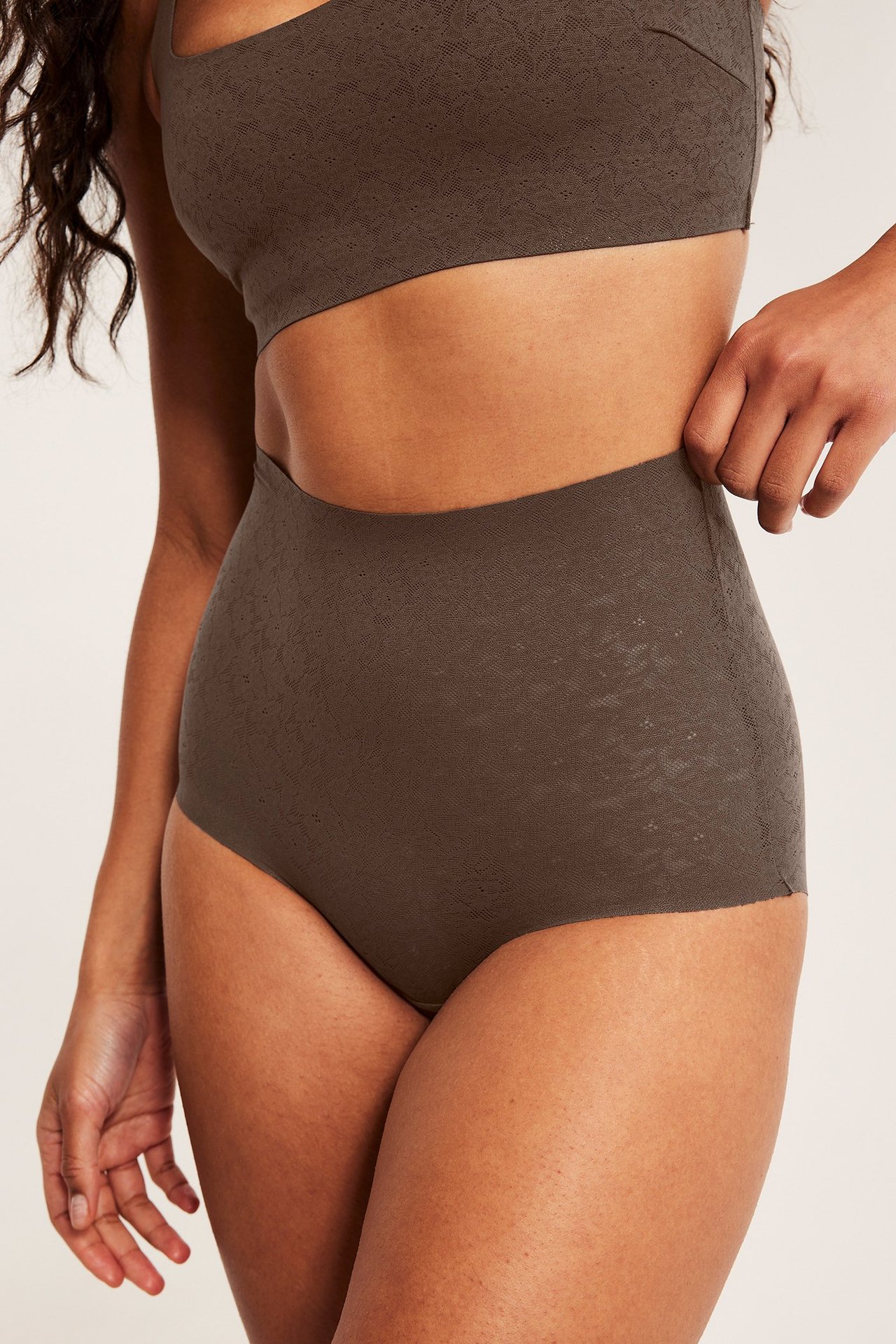 Lähikuva naisesta tummanruskeissa korkeavyötäröisissä pitsihipstereissä ja yhteensopivassa rintaliivissä.