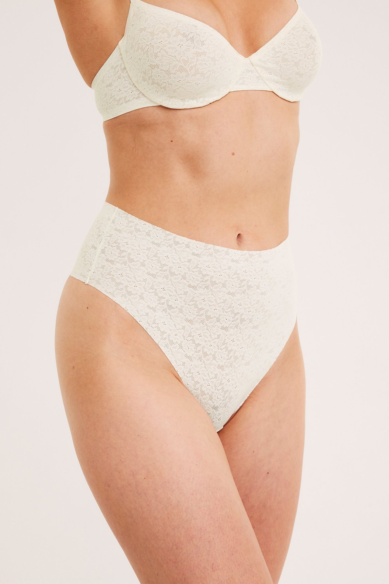 Front-sidevisning av en kvinne i offwhite høy midje blondestringtruse og matchende blonde-BH.