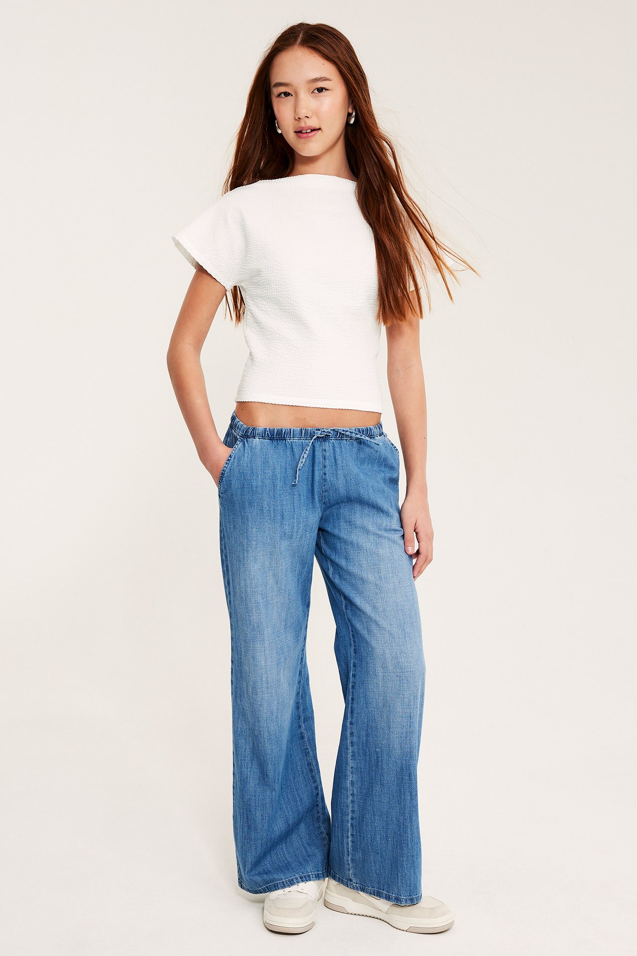 Framifrån: Barn i vit båtringad fladdermusärmstopp, ljusblå vida byxor med dragsko och sneakers.