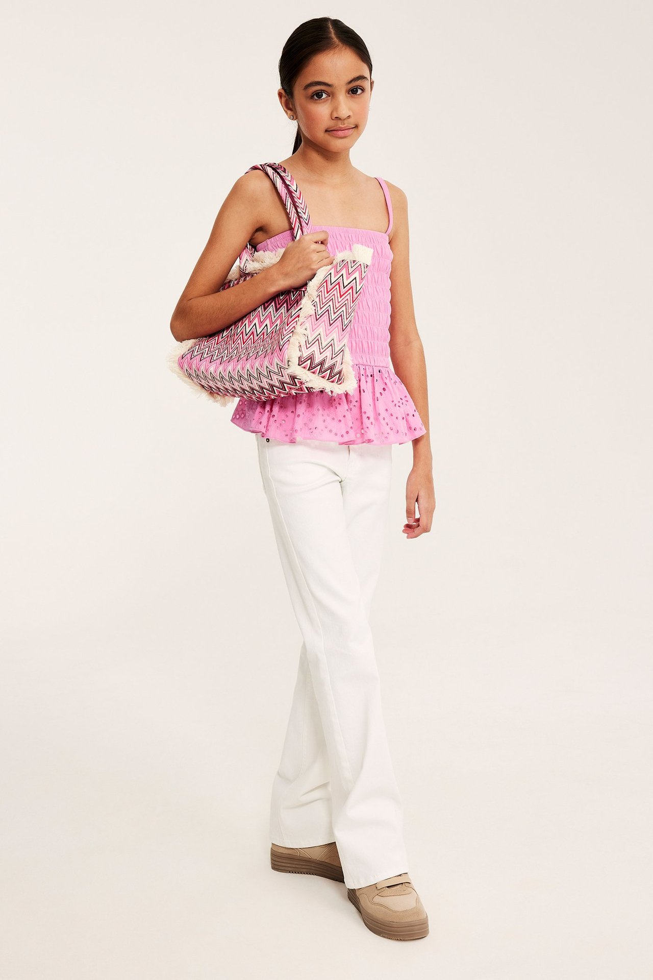 Edestä: Lapsi vaaleanpunaisessa smokkitopissa brodeeratulla röyhelöllä, valkoiset housut, kuviollinen kassi.