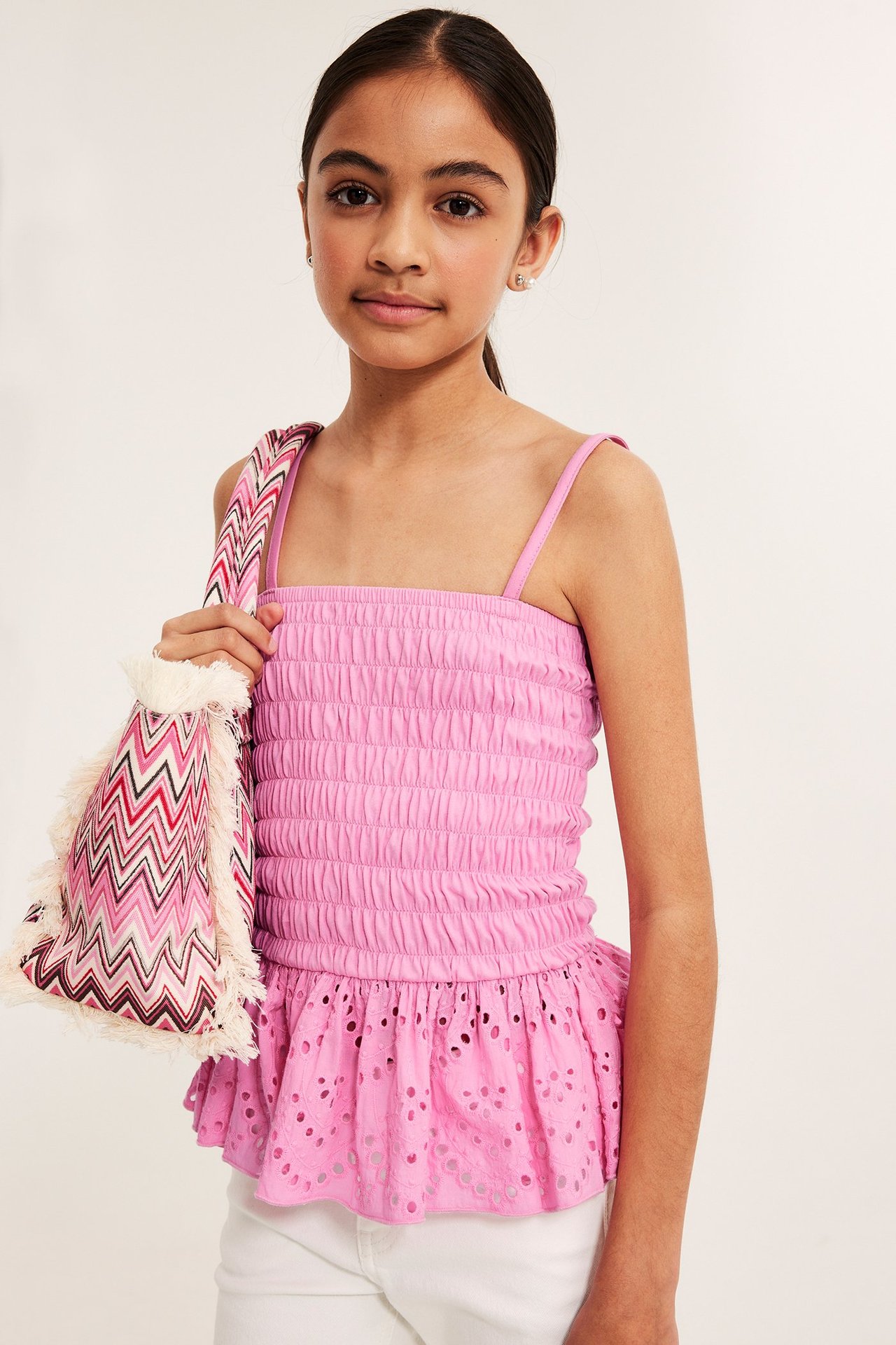 Widok z przodu: Dziecko w różowym marszczonym topie z falbanką i haftem, trzymające wzorzystą torbę.