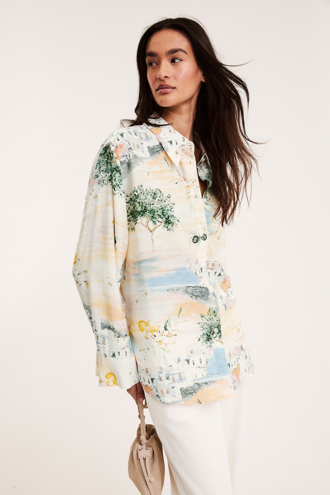 Front view: Kvinna i lös offwhite mönstrad viskosskjorta, vita byxor, håller beige väska.