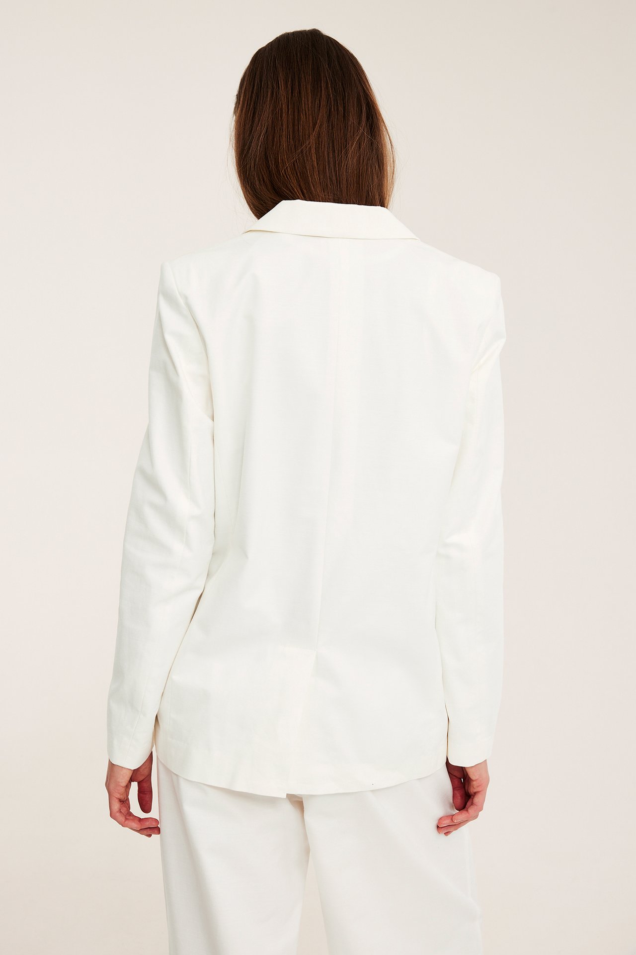 Widok od tyłu: kobieta w białej oversizowej marynarce z mieszanki bawełny na białych spodniach.