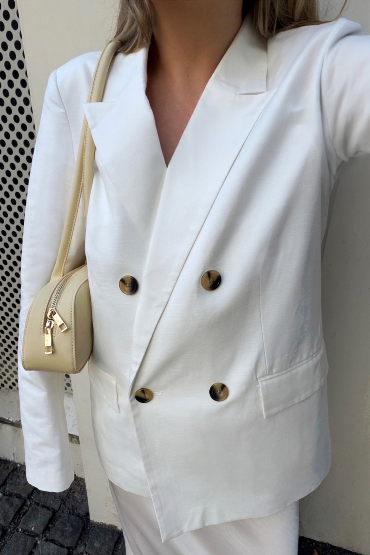 Zbliżenie na kobietę w białej, oversize'owej dwurzędowej marynarce z brązowymi guzikami, niosącą beżową torebkę.