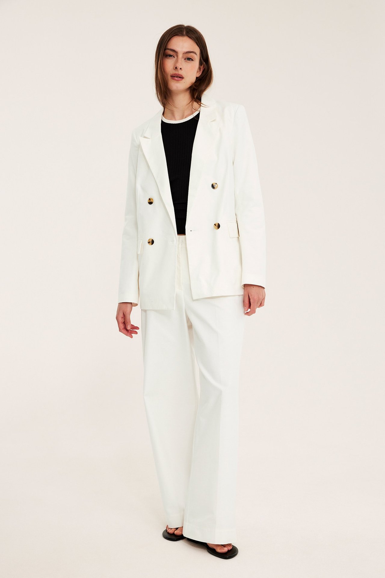 Widok z przodu: Kobieta w białej, dwurzędowej marynarce oversize ze złotymi guzikami, czarnym topie i białych spodniach.