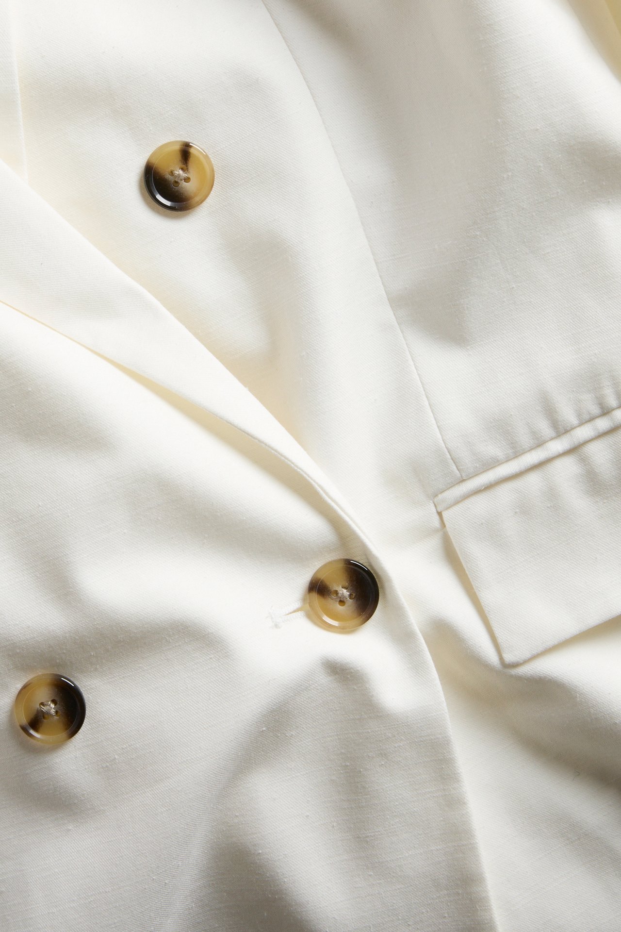 Nærbilde av en hvit dobbeltspent blazer med brune plasthornknapper og en frontlomme.