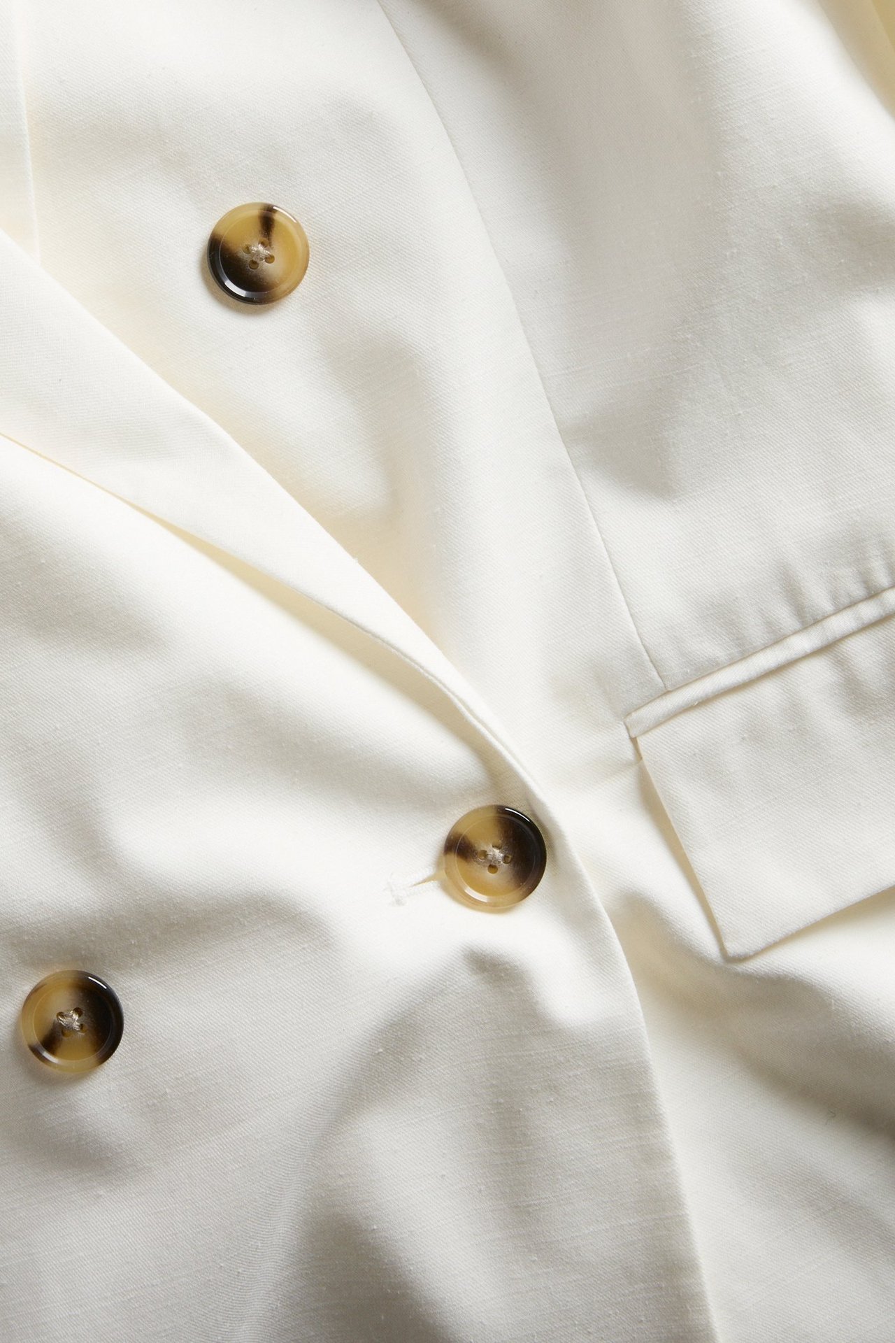 Nærbilde av en hvit dobbeltspent blazer med brune plasthornknapper og en frontlomme.