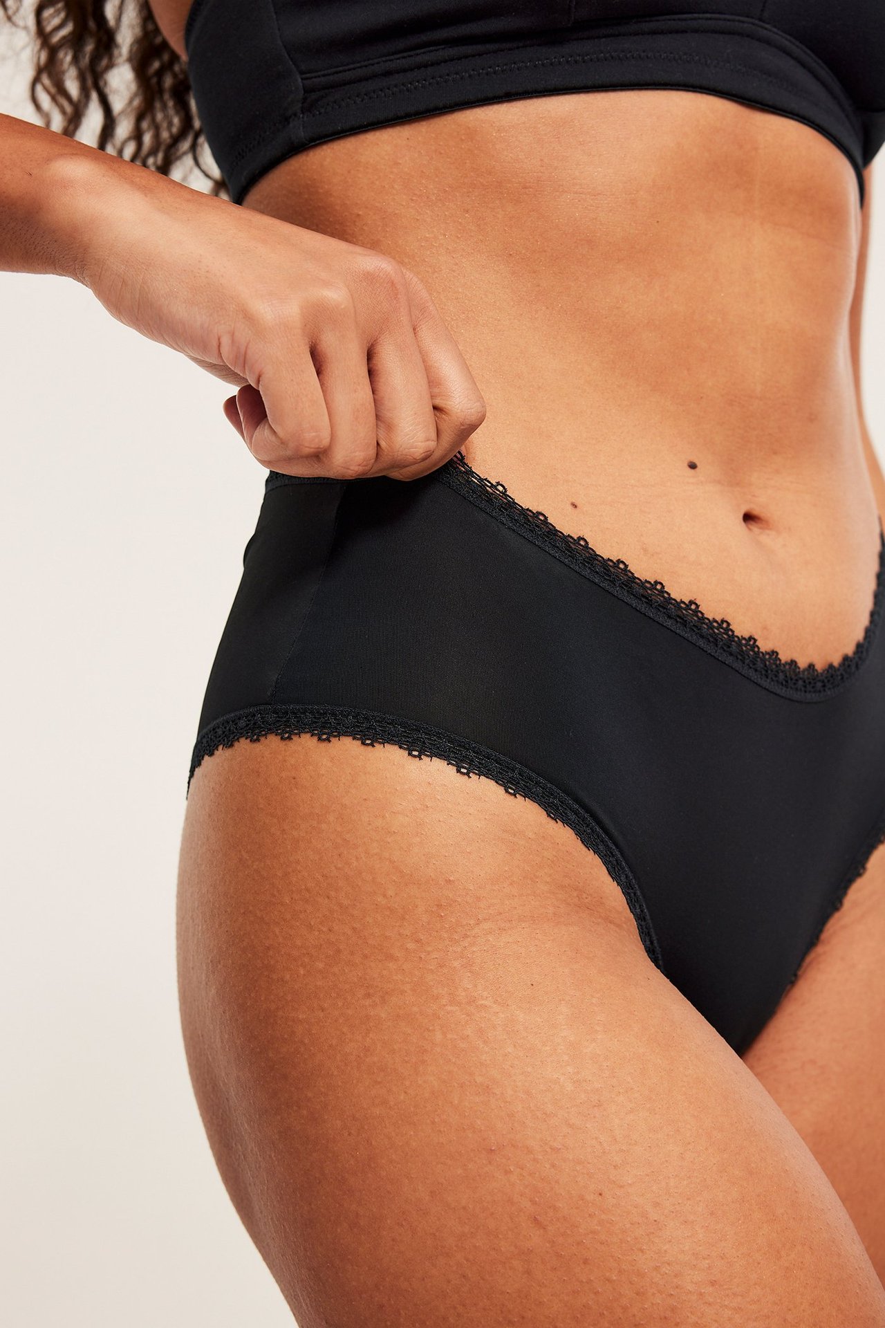 Närbild framifrån på kvinna i svarta mikro cheeky trosor med spetskant, drar i linningen, och svart BH.