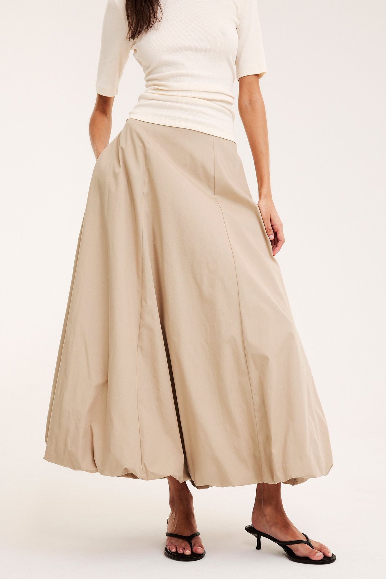 Edestä: Nainen beigessä puuvillapallohameessa, valkoisessa topissa ja mustissa korollisissa varvassandaaleissa.