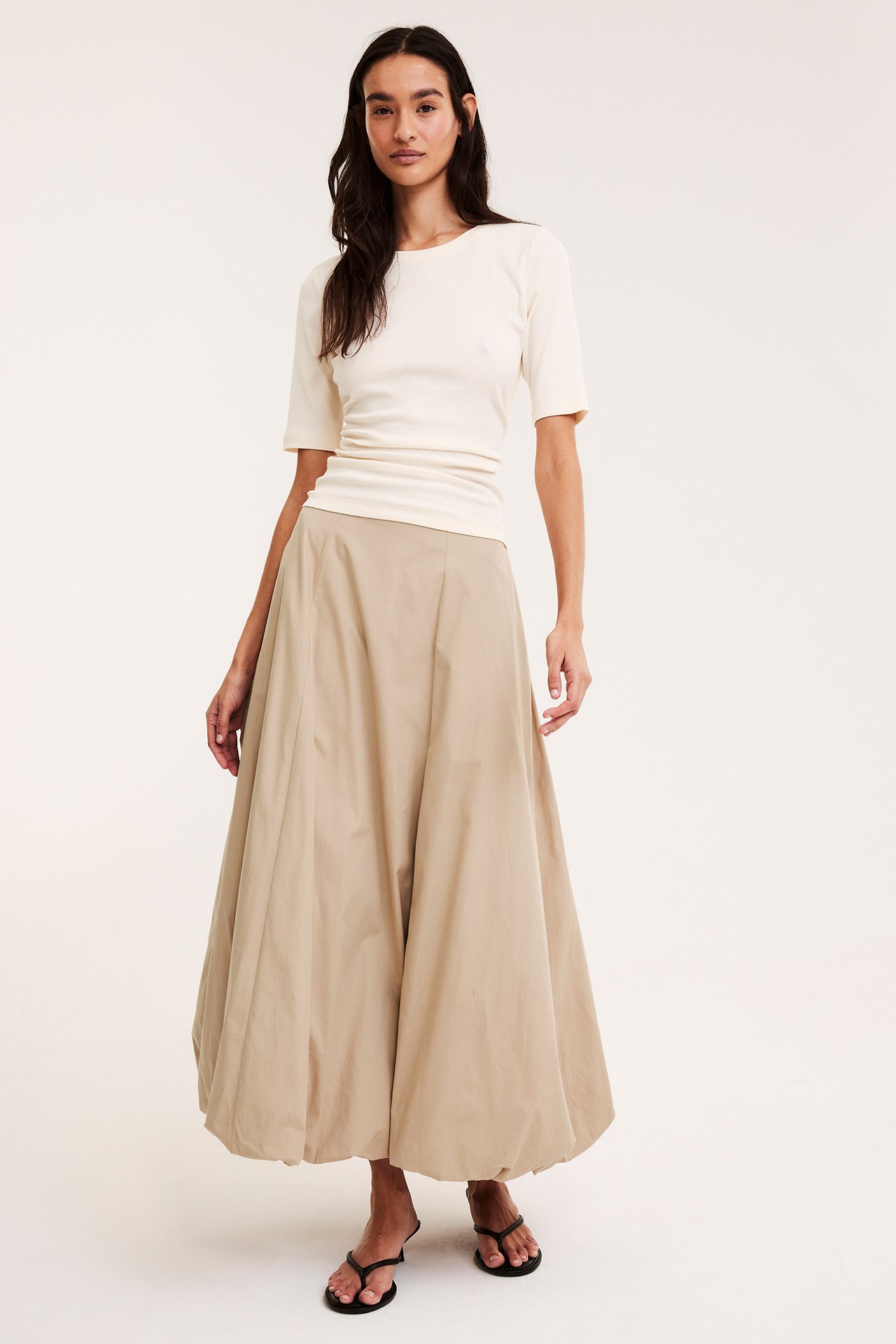 Frontvy: Kvinna i lång beige ballongkjol i bomullspoplin, gräddvit ribbad topp och svarta sandaler.