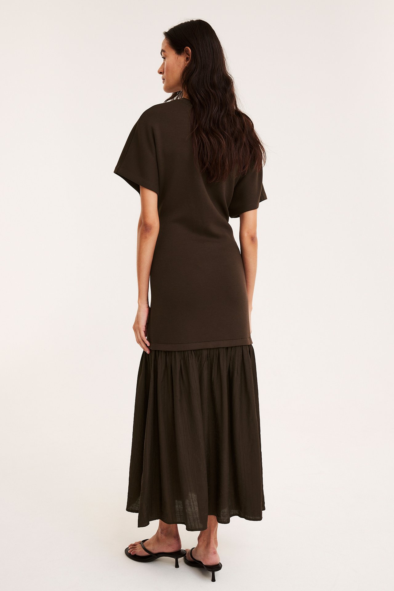 Kobieta tyłem w ciemnobrązowej długiej sukience z dzianiny i tkaniny, z czarnymi sandałami na obcasie.