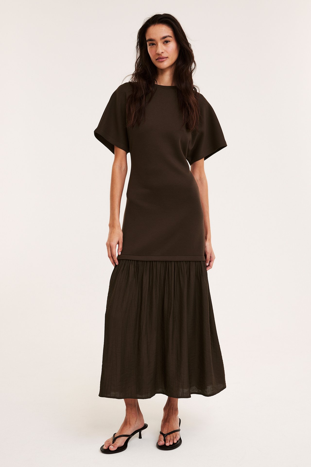 Framifrån: kvinna i mörkbrun långklänning med jerseytopp, vävd kjol och svarta klacksandaler.