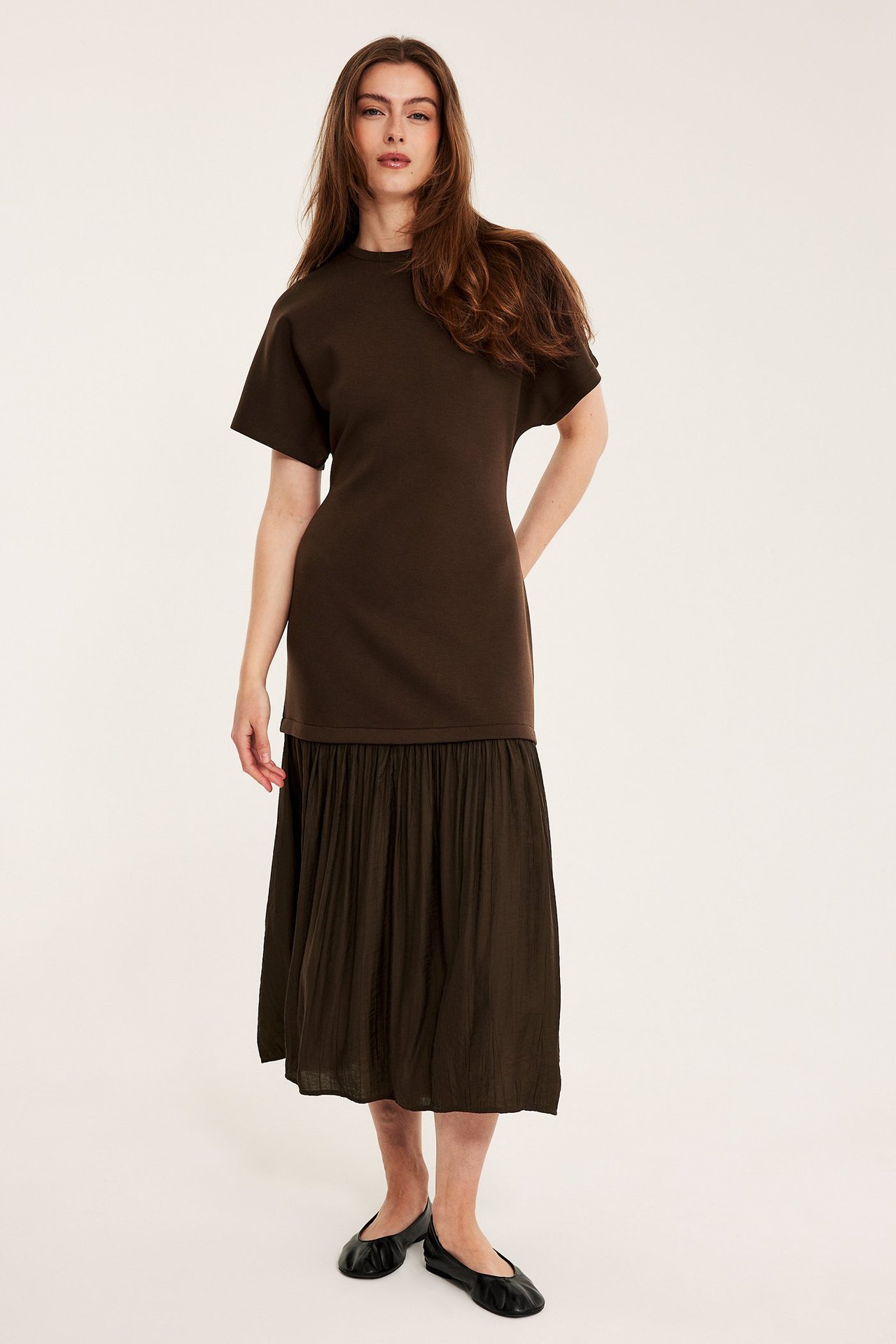 Edestäpäin: nainen tummanruskeassa pitkässä mekossa, jerseypaita, kudottu hame, ja mustat ballerinat.