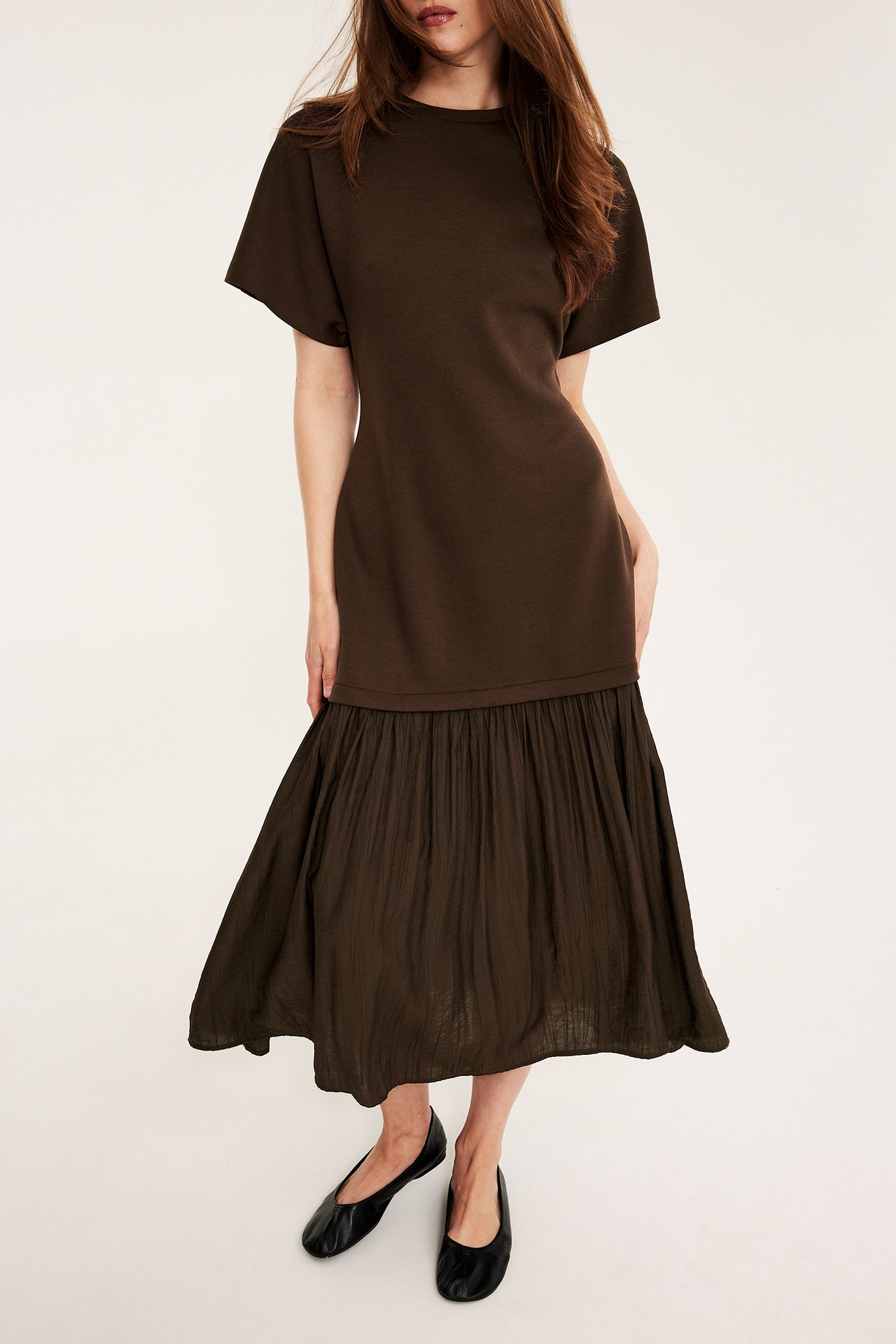 Edestä: Nainen tummanruskeassa pitkässä mekossa, jossa trikoo-yläosa ja kudottu hame, mustat ballerinat.