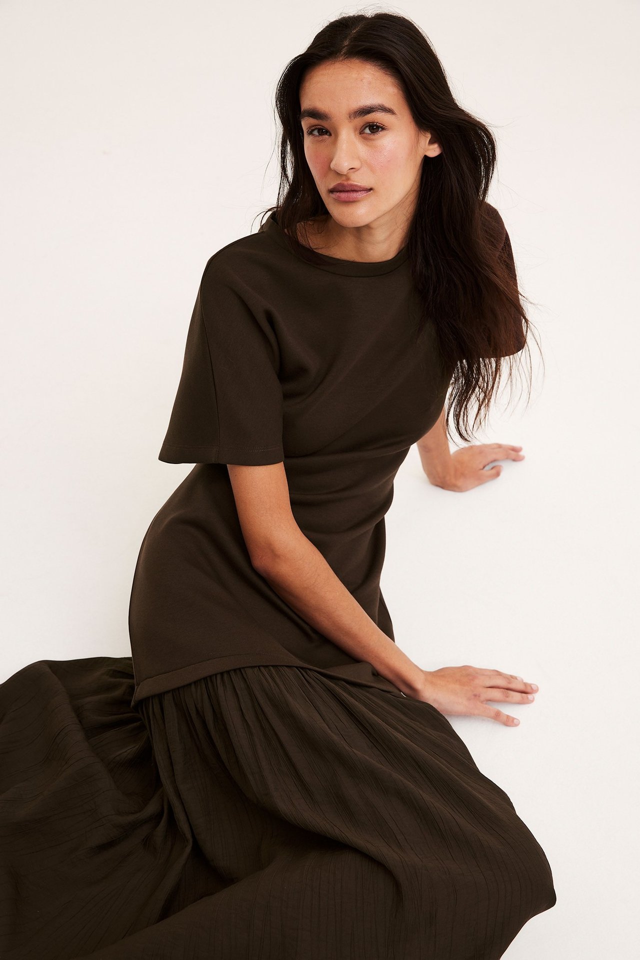 Widok z przodu kobiety w ciemnobrązowej długiej sukience z dżersejową górą, krótkim rękawem i tkaną spódnicą.