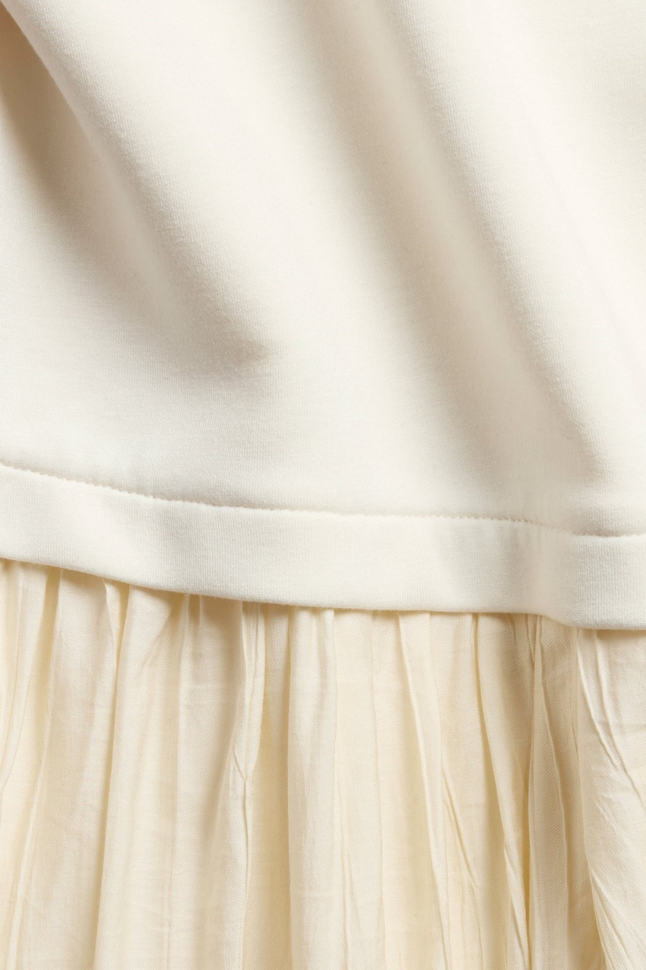 Nærbilde av en kremhvit lang kjole med glatt jerseytop som går over i et plissert vevd skjørt.