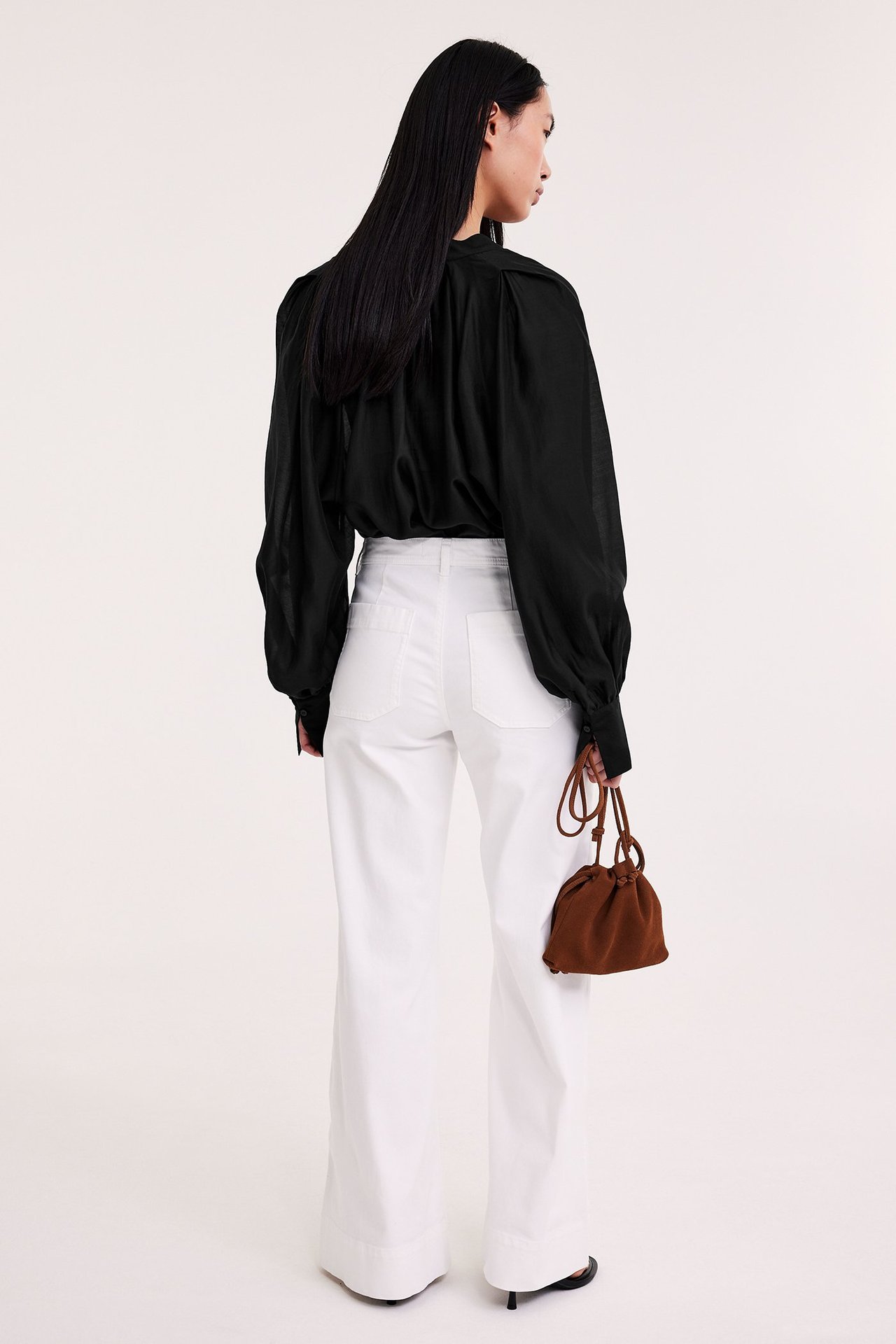 Ryggbilde av kvinne i svart voluminøs bluse, hvite vide jeans, holder en liten brun snøreveske.