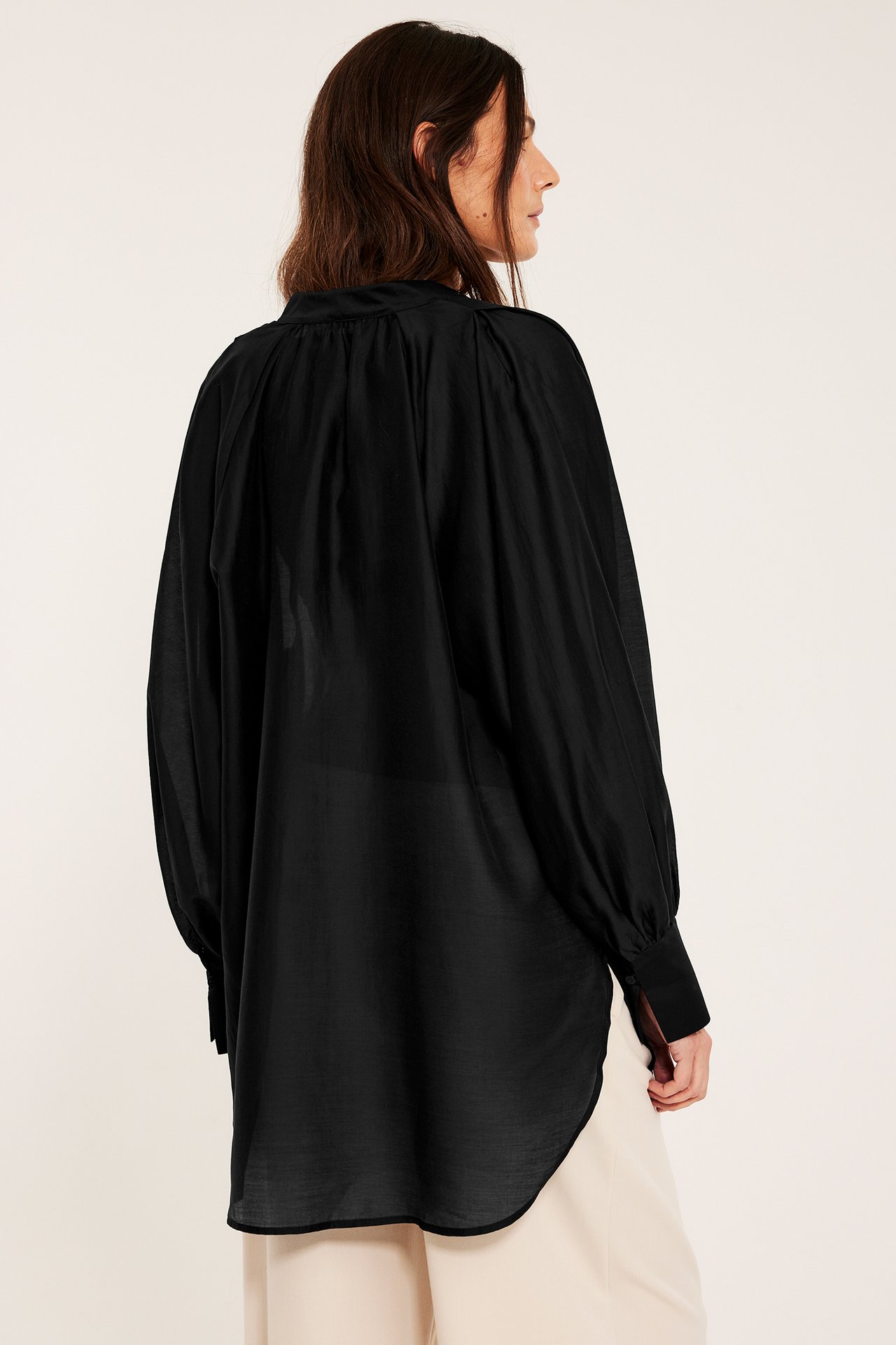 Bakfra/siden av kvinne i voluminøs svart bluse med rynkede ermer og lys beige bukse.