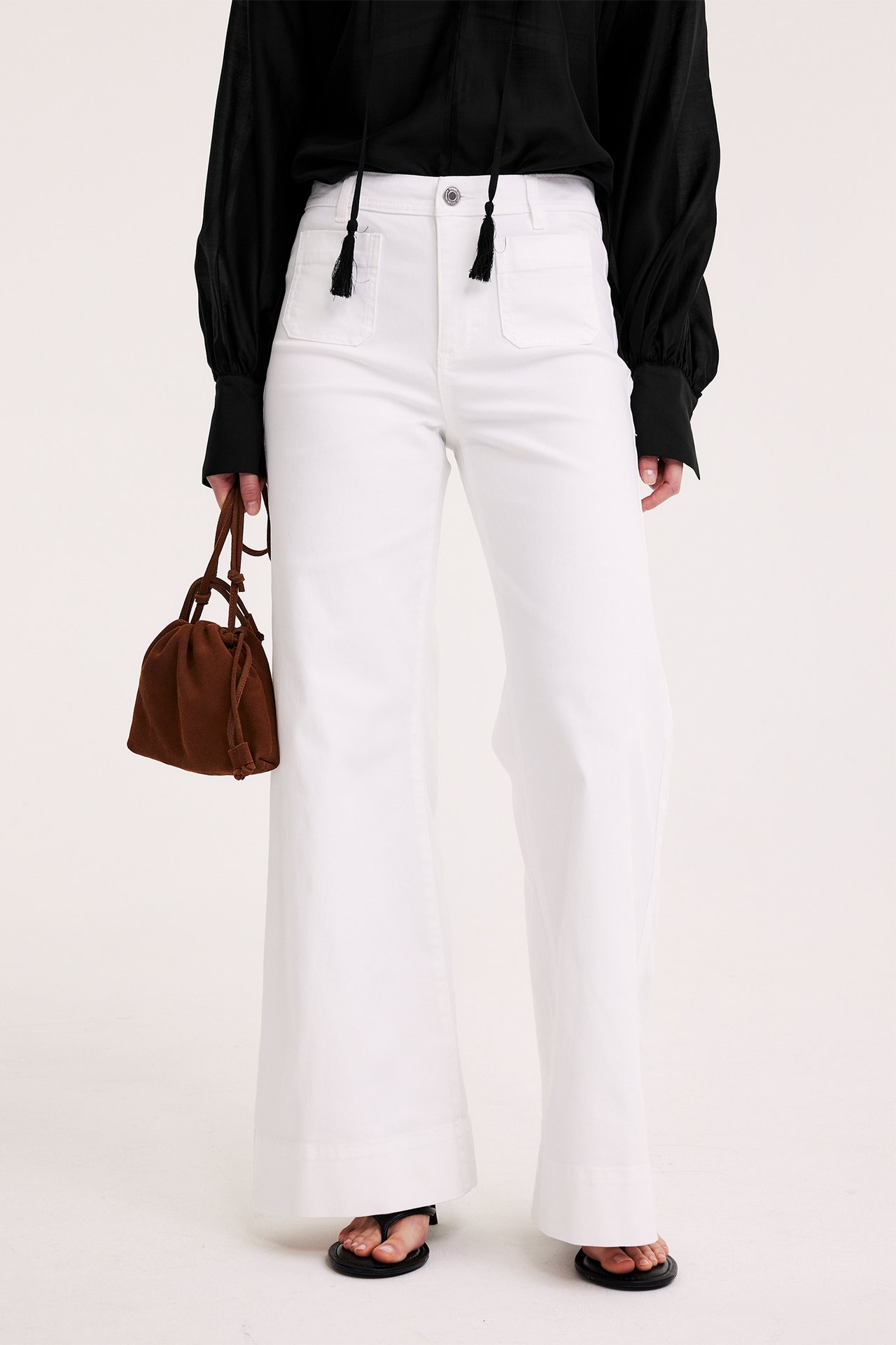 Forfra: Kvinne i svart voluminøs bluse, hvite vide bukser, holder brun semsket veske.