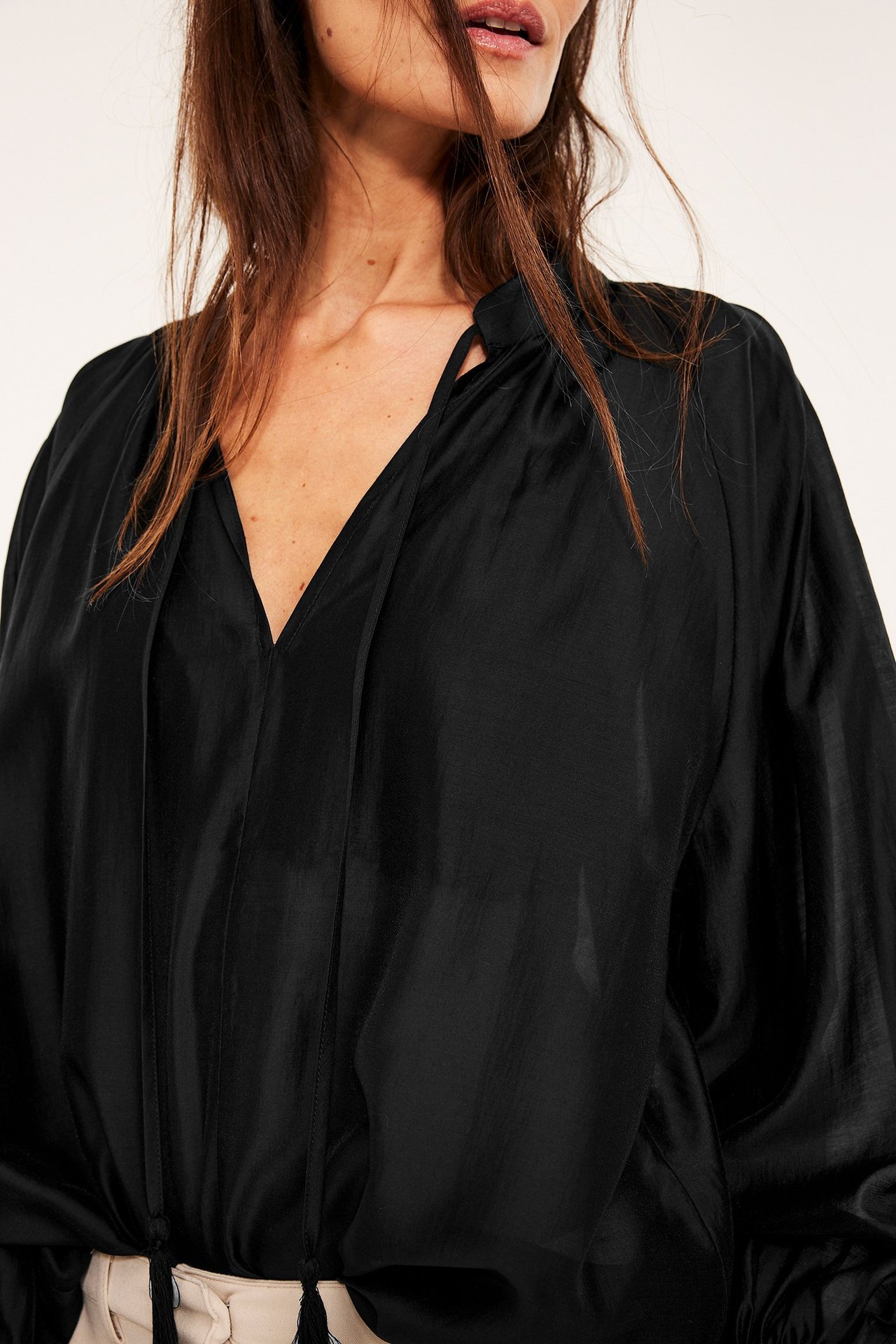 Frontbilde: Kvinne i svart voluminøs lyocellblandingsbluse med V-hals og knytebånd, over lyse bukser.