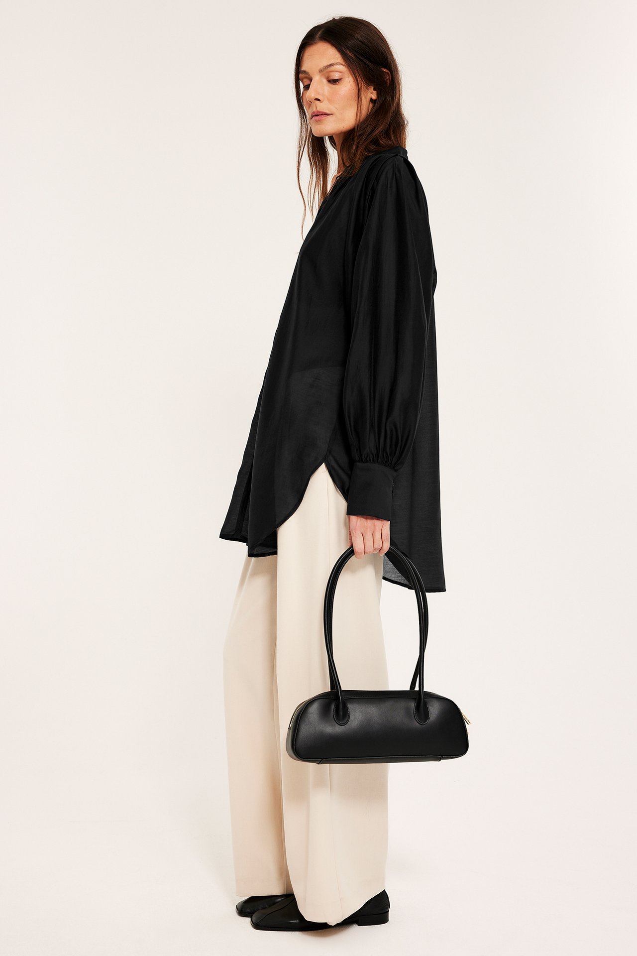 Sidovy av kvinna i svart voluminös blus, beiga vida byxor och svarta skor, håller svart handväska.