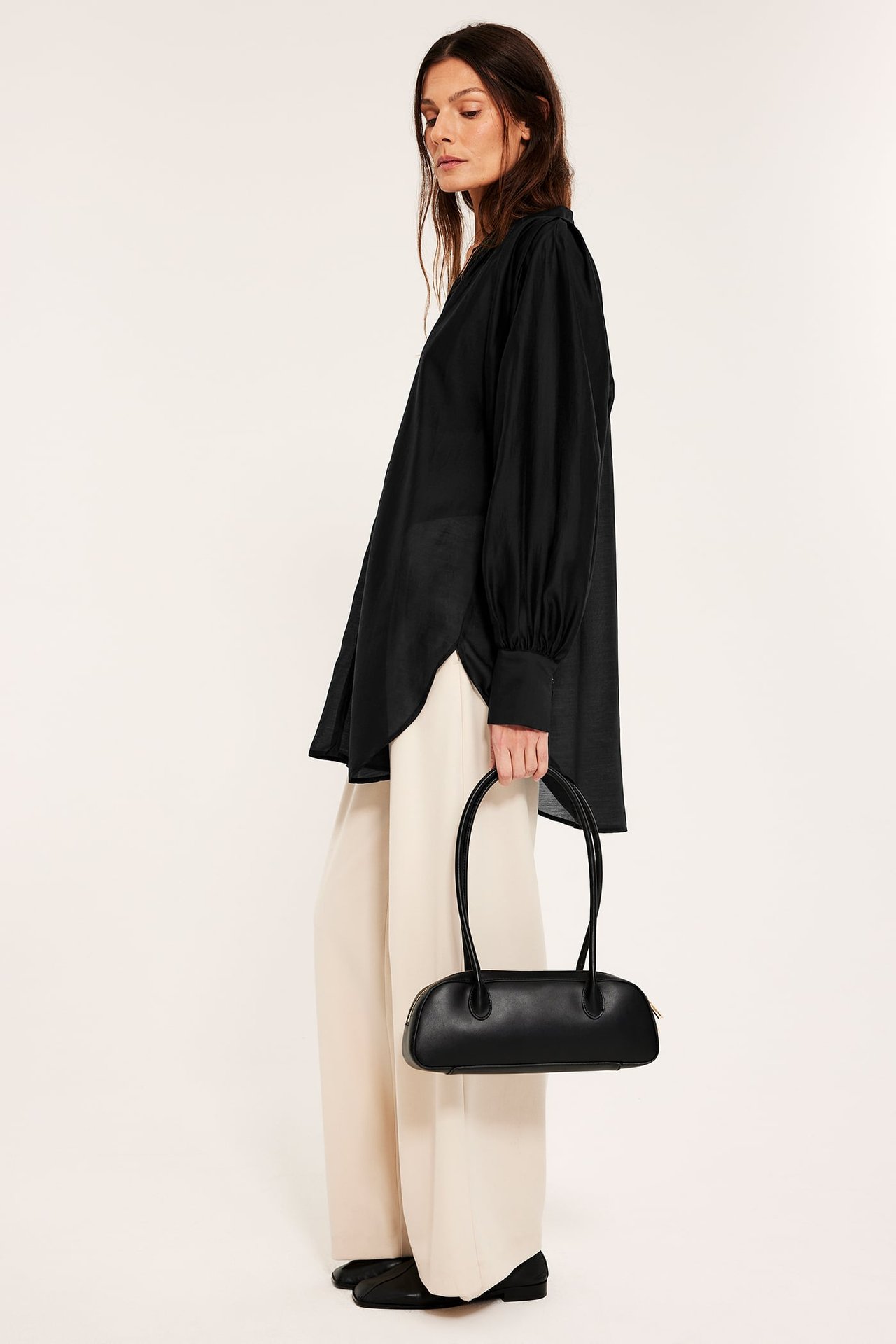Sivukuva naisesta mustassa volyymipuserossa, beigissä leveissä housuissa ja kengissä, pitäen mustaa käsilaukkua.