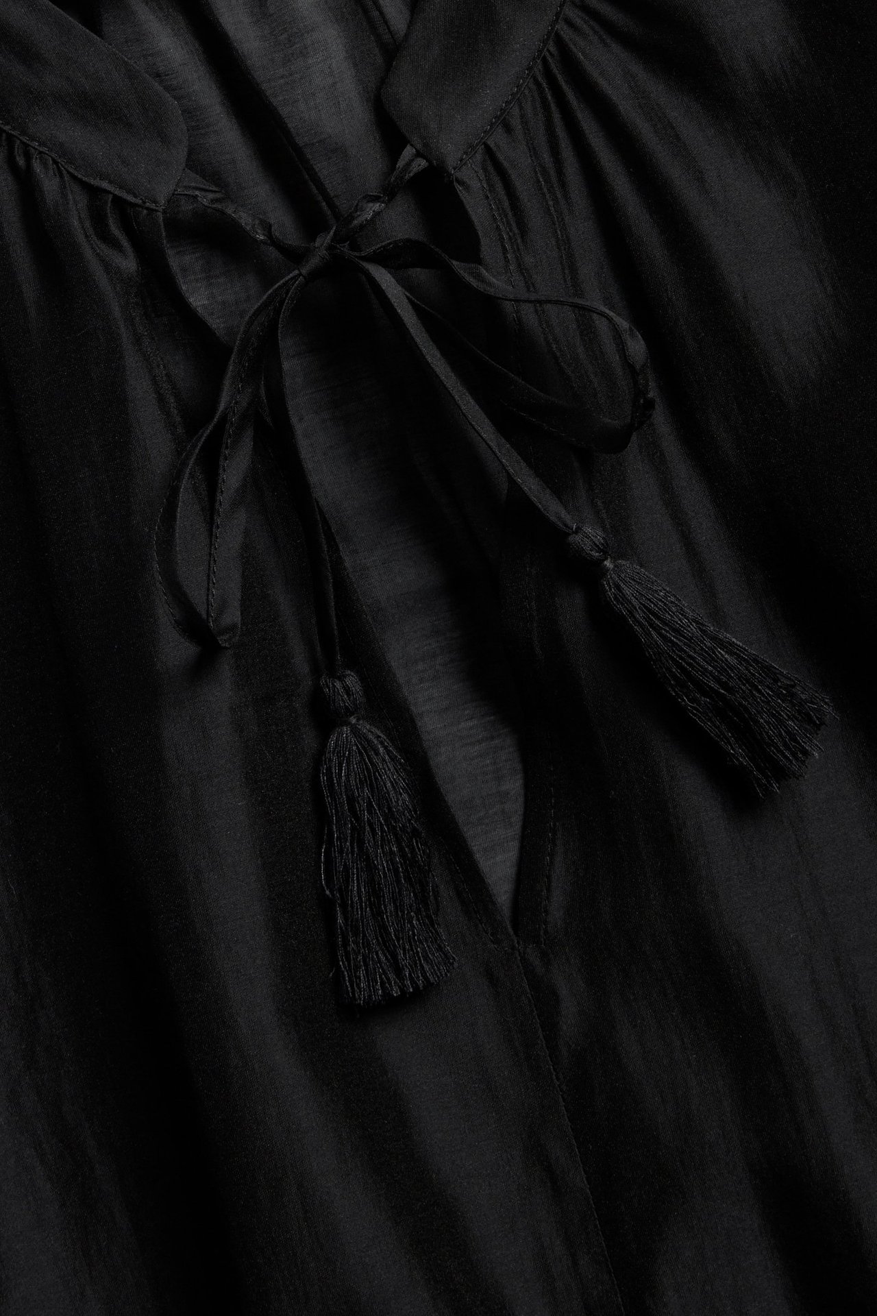 Nærbilde av en svart voluminøs bluse med V-hals og knytebånd med dusker.