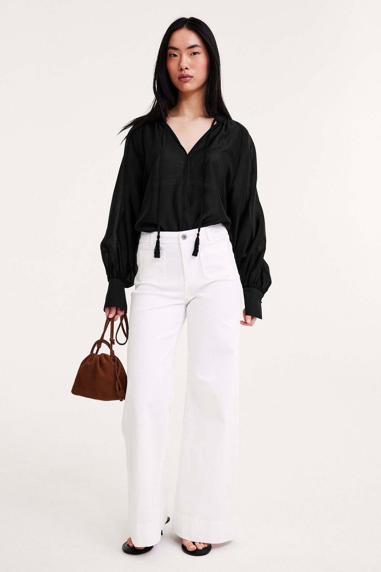 Frontvisning: kvinne i svart voluminøs bluse med knytebånd, hvite vide bukser, holder brun bøtteveske.