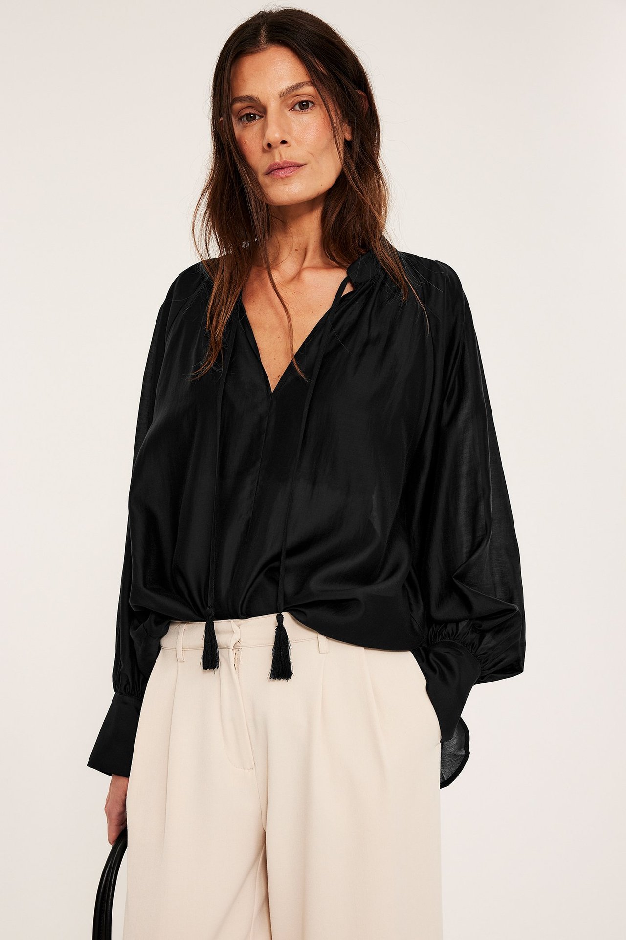 Widok z przodu: kobieta w czarnej obszernej bluzce z wiązaniami i jasnobeżowych szerokich spodniach.