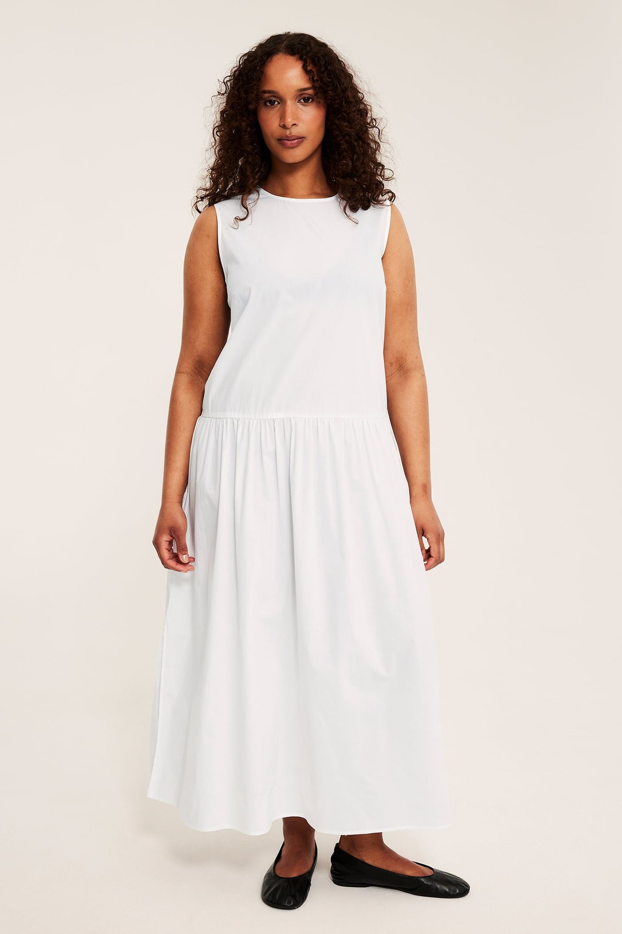 Framifrån: kvinna i en löst sittande vit ärmlös bomullsklänning och svarta ballerinaskor.