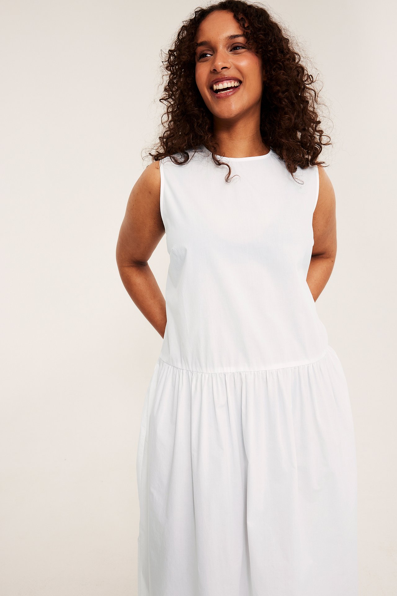 Frontvy av en leende kvinna i en vit, löst sittande, ärmlös bomullsklänning med rynkad midja.