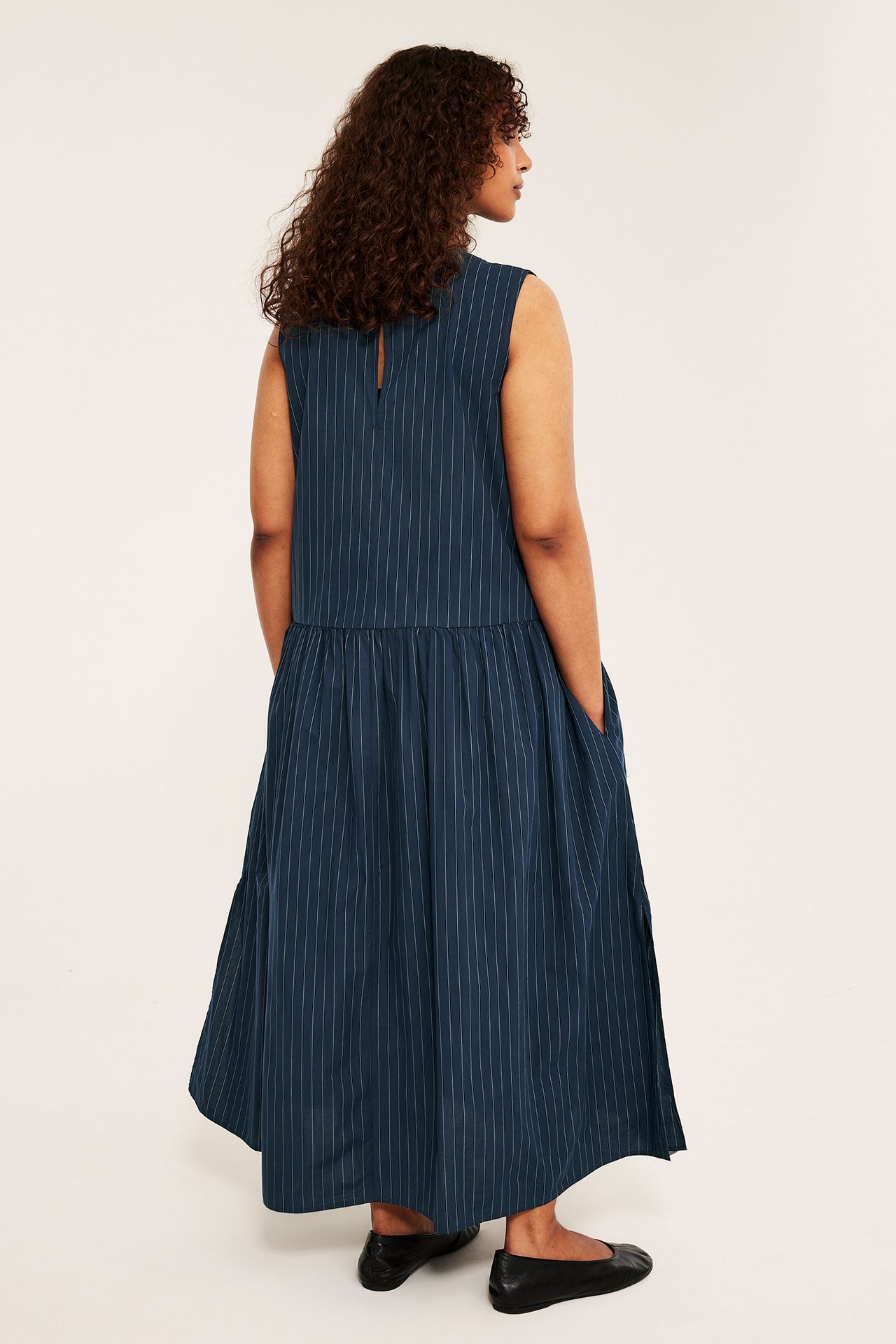 Widok z tyłu kobiety w granatowej, luźnej, bezrękawnej bawełnianej sukience maxi w paski i czarnych baletkach.