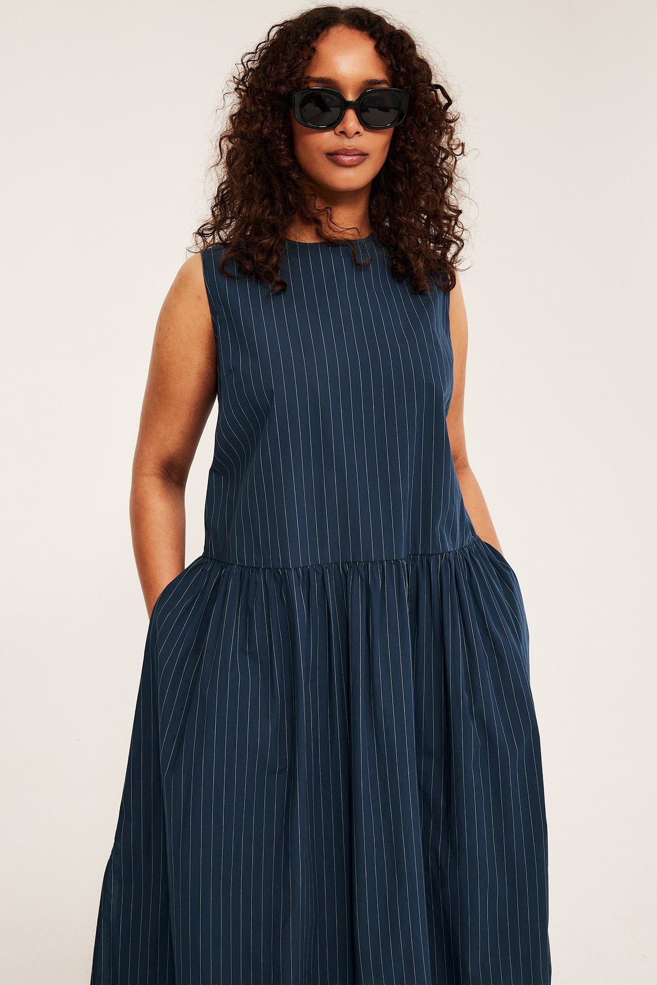 Framifrån, kvinna i mörkblå randig ärmlös bomullsklänning med fickor, bär svarta solglasögon.