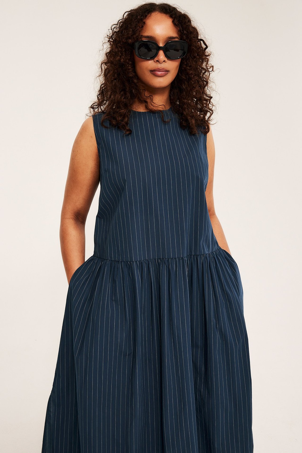Forfra, kvinne i mørkblå, stripet ermeløs bomullskjole med lommer, bærer svarte solbriller.
