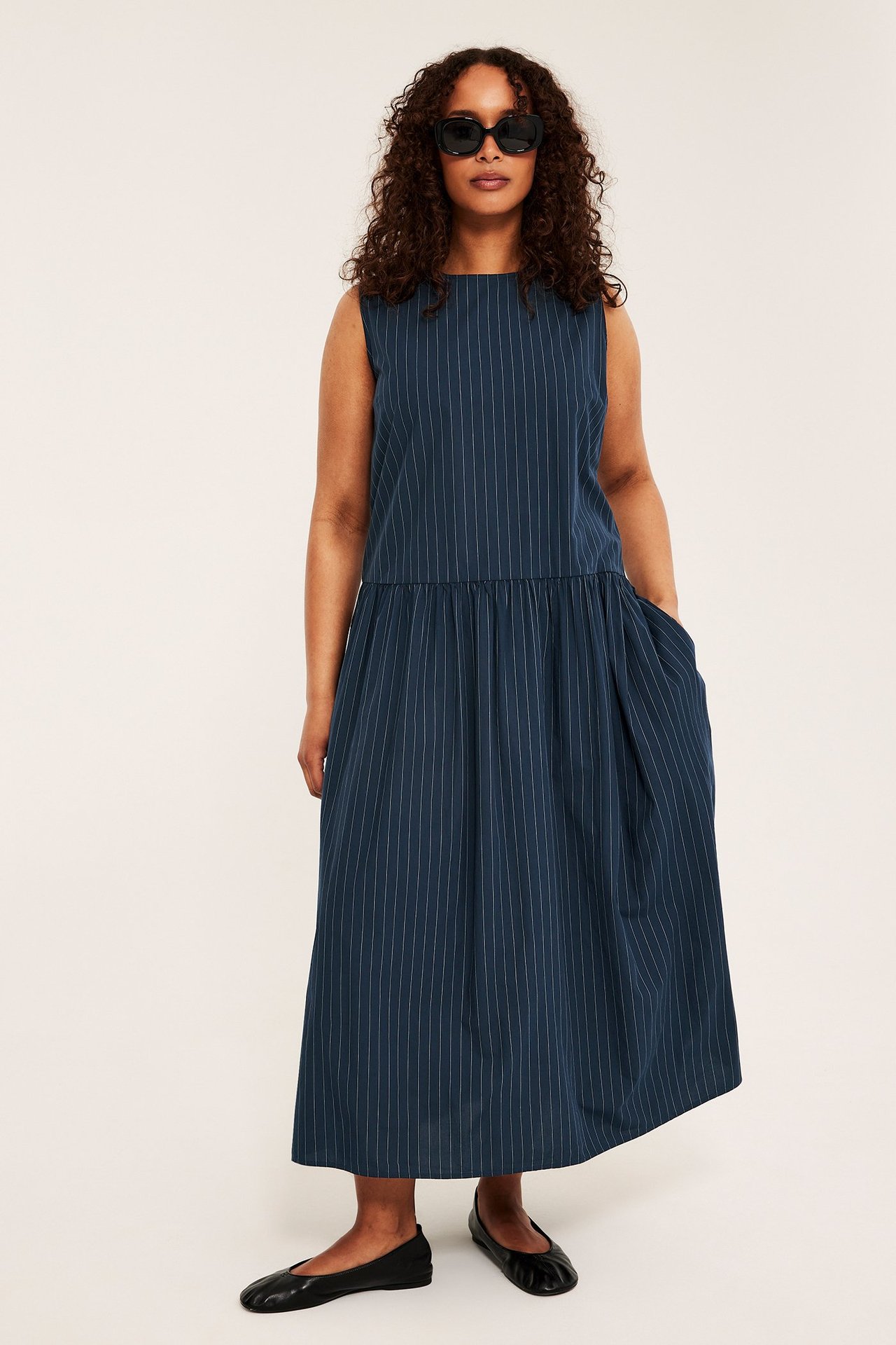 Kobieta w granatowej sukience w paski, bez rękawów, z czarnymi okularami i baletkami, widok z przodu.